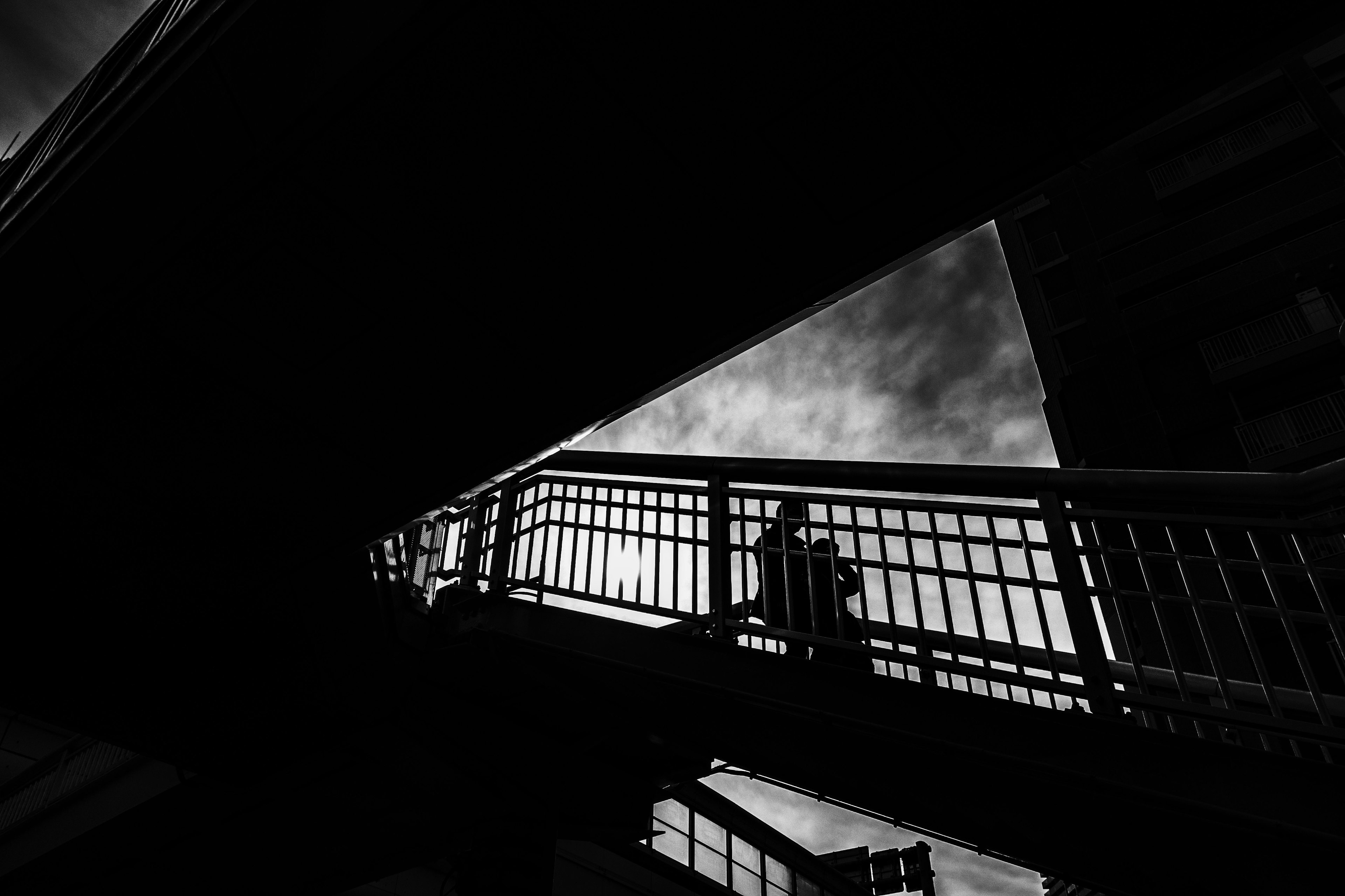 High-contrast black and white urban scene with a pedestrian walking on a bridge structure sharp angles and a glimpse of the sky