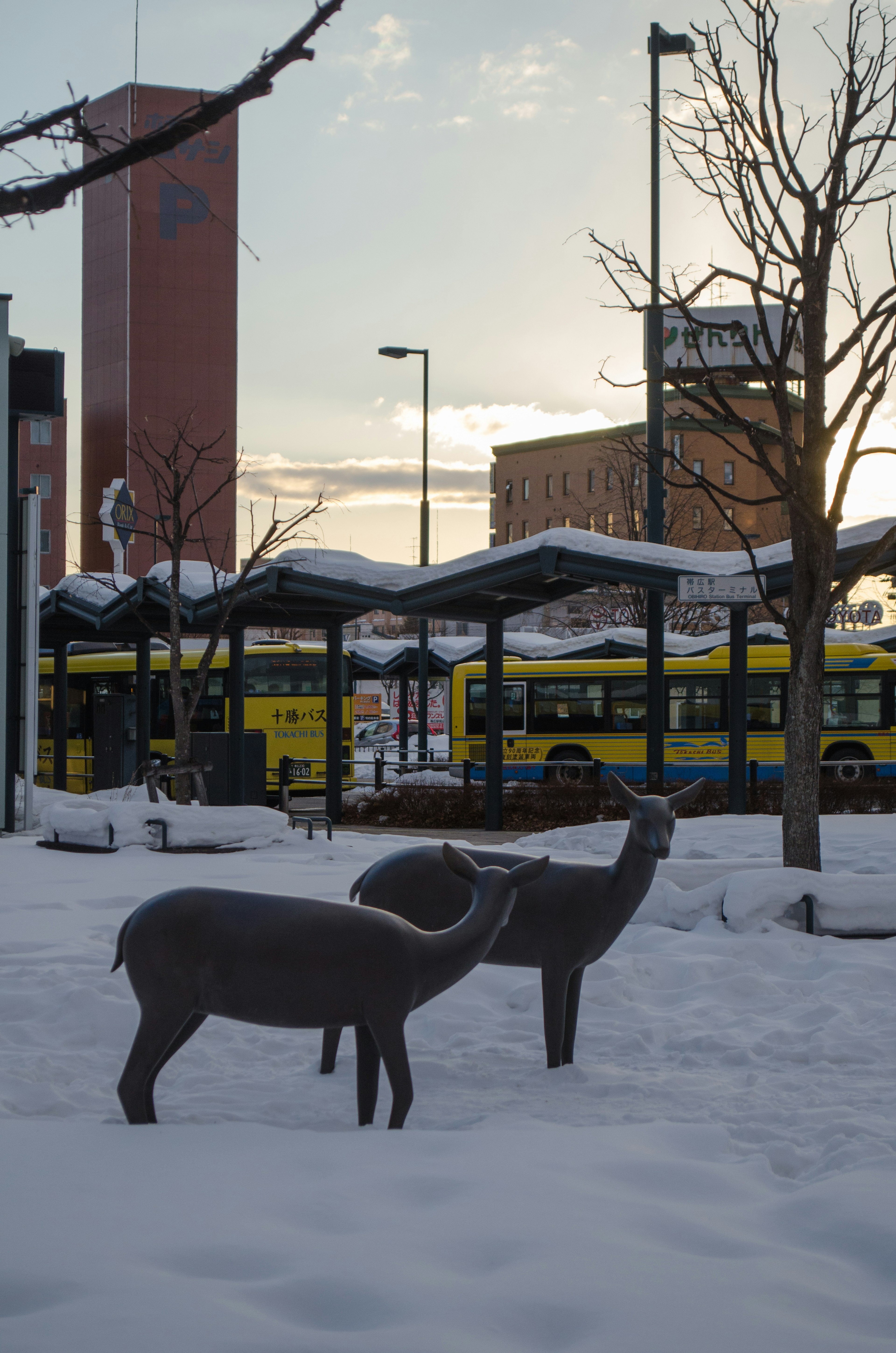 Deer sculptures standing in snow with a bus station in the background