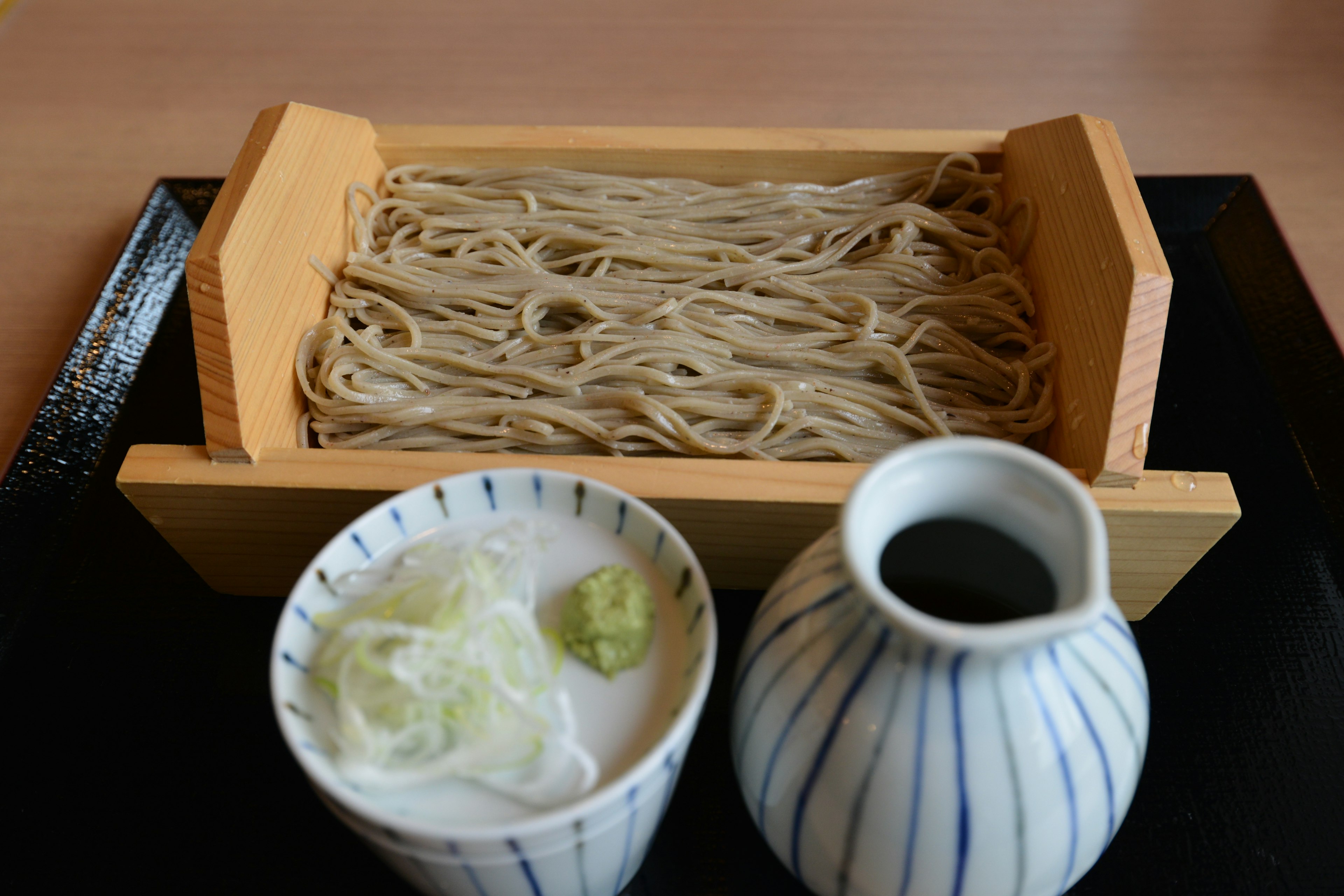 Soba noodles served in a wooden tray with a bowl of condiments and a ceramic sauce pitcher