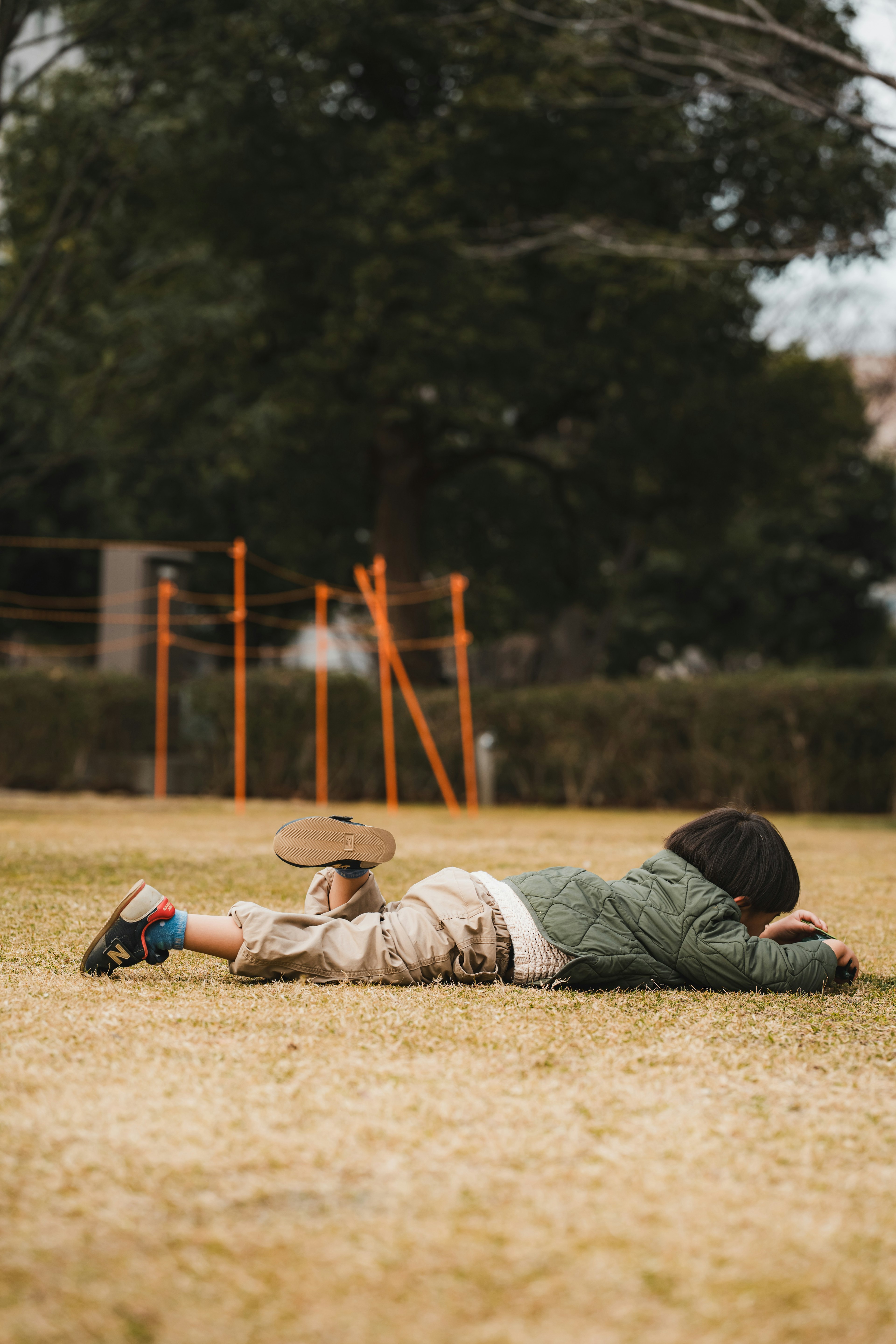 Child lying on grass in a park wearing a green jacket and beige pants