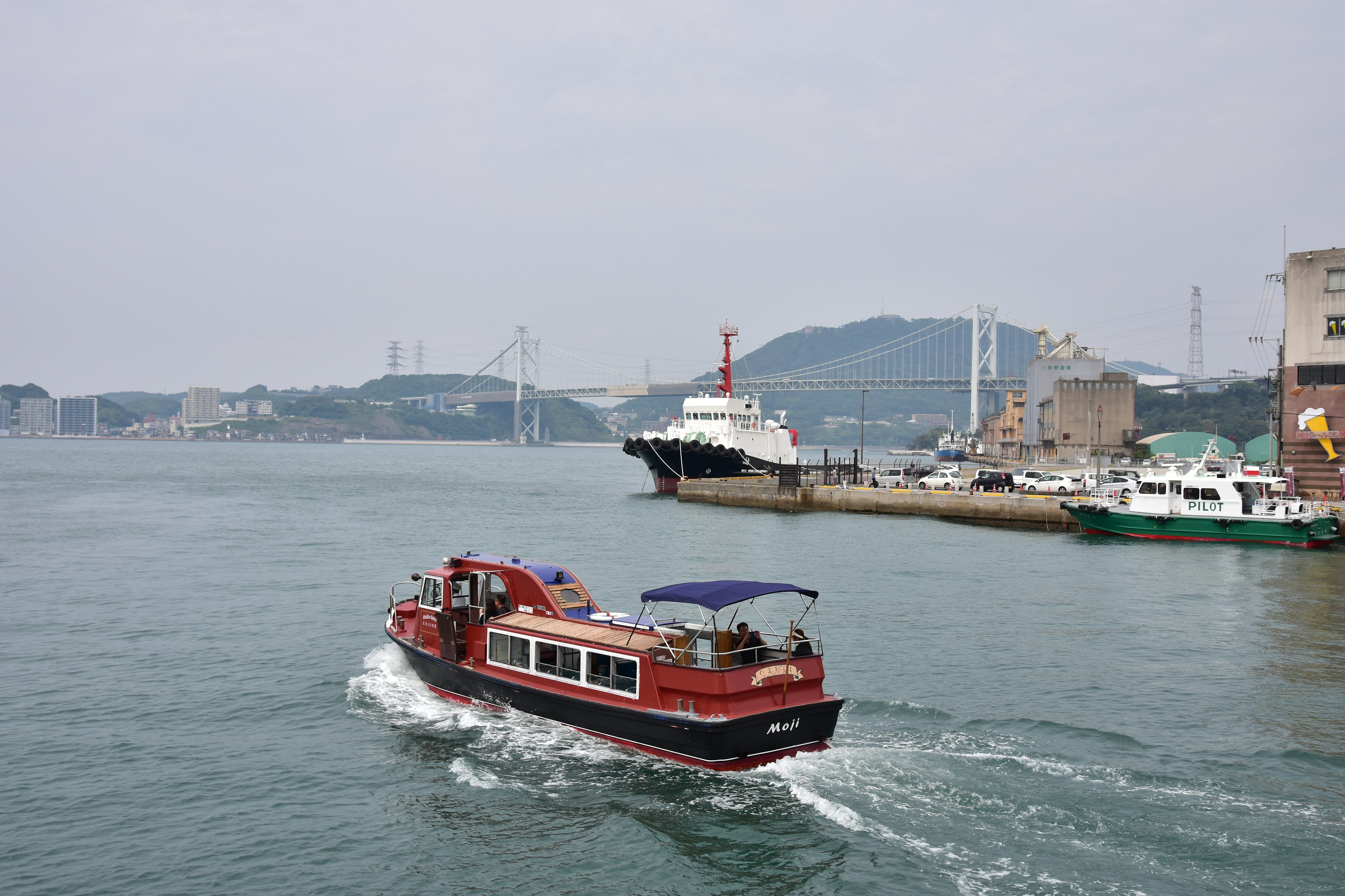 Ein rotes Boot gleitet über das Wasser mit einem Hafen und Bergen im Hintergrund