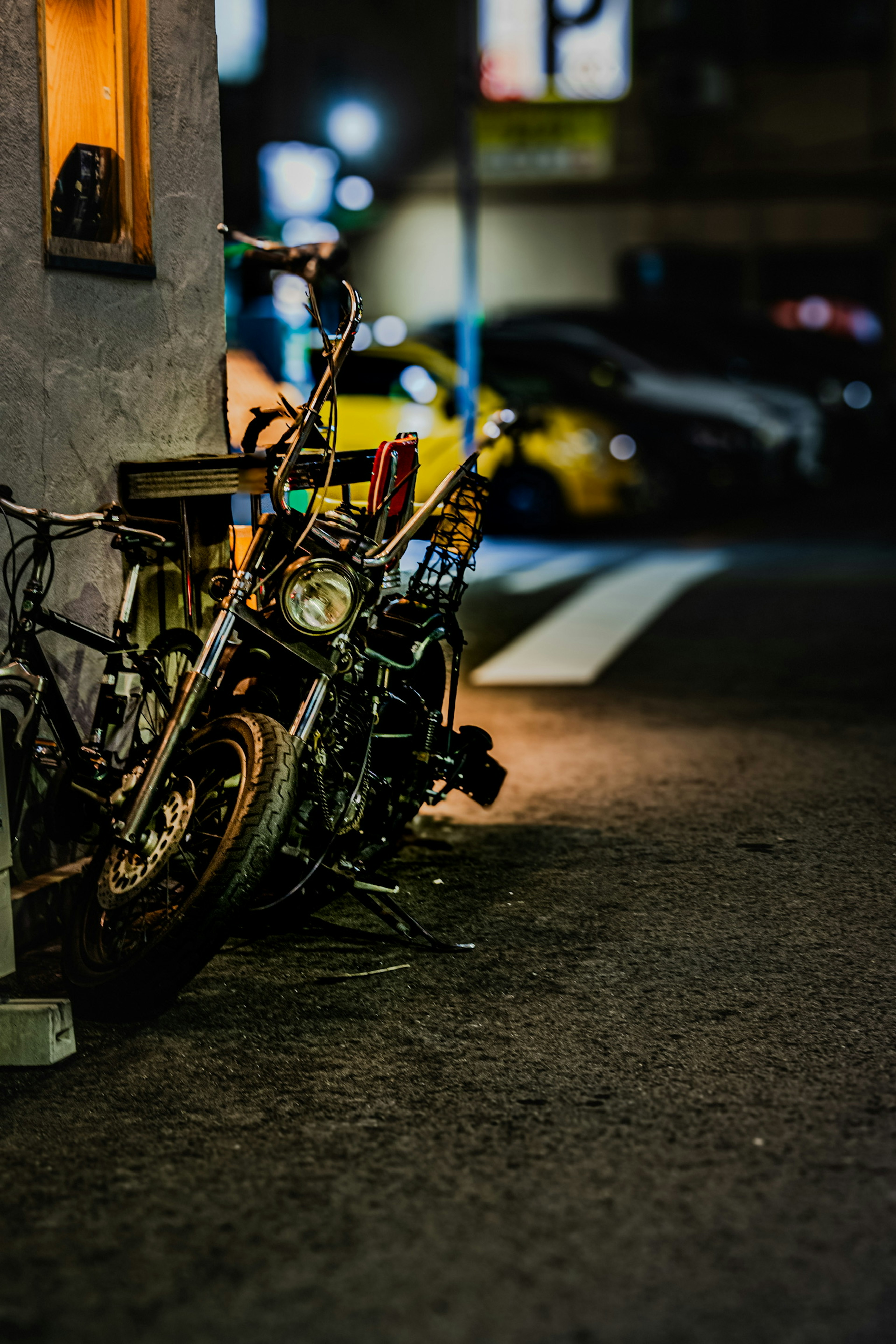 Ein altes Motorrad, das an einer Wand in einer schwach beleuchteten Gasse lehnt, mit verschwommenen Lichtern im Hintergrund