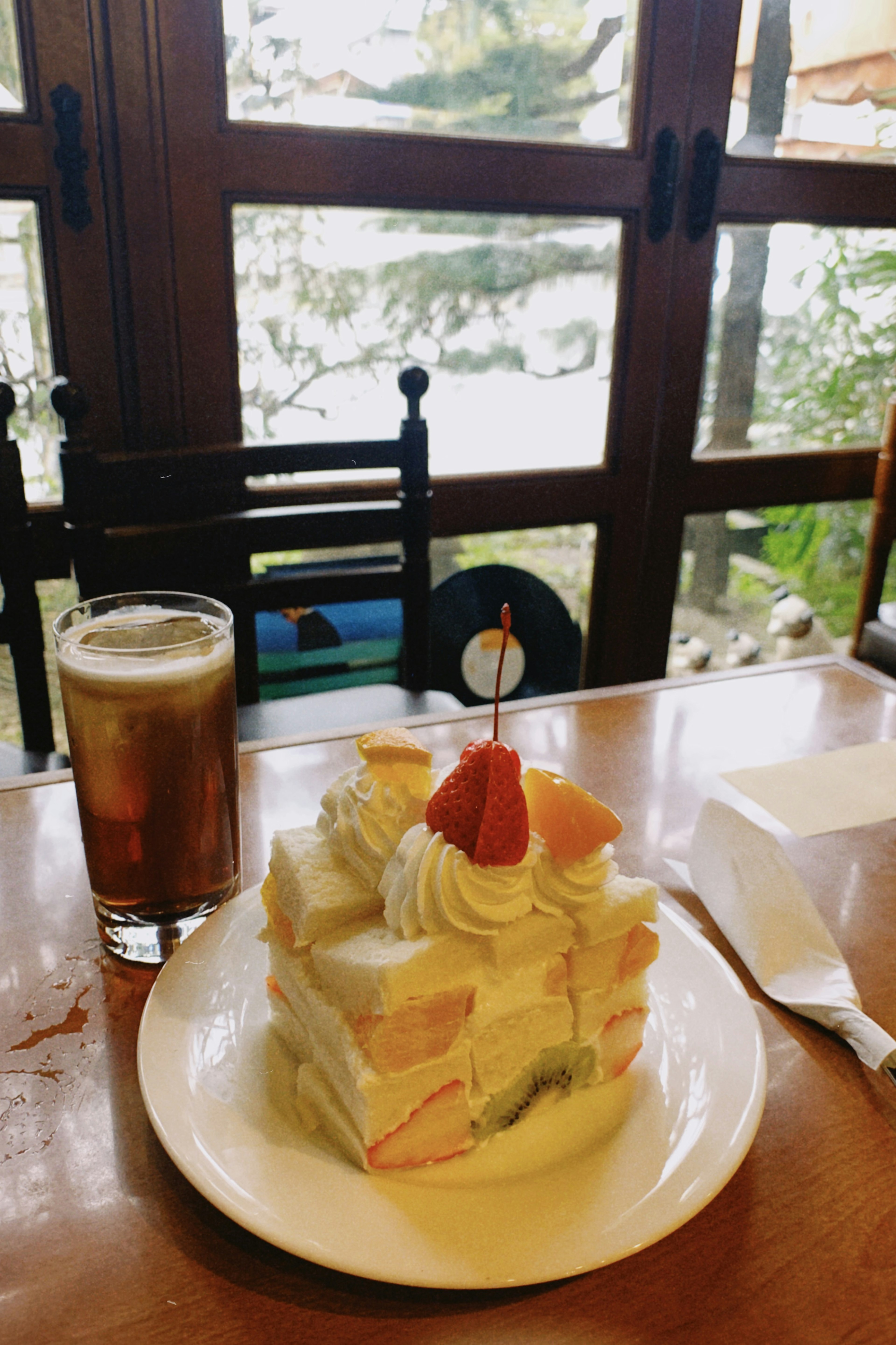 Ein Obstkuchen mit Schlagsahne und einer Kirsche obendrauf neben einem Glas Eiskaffee auf einem Holztisch