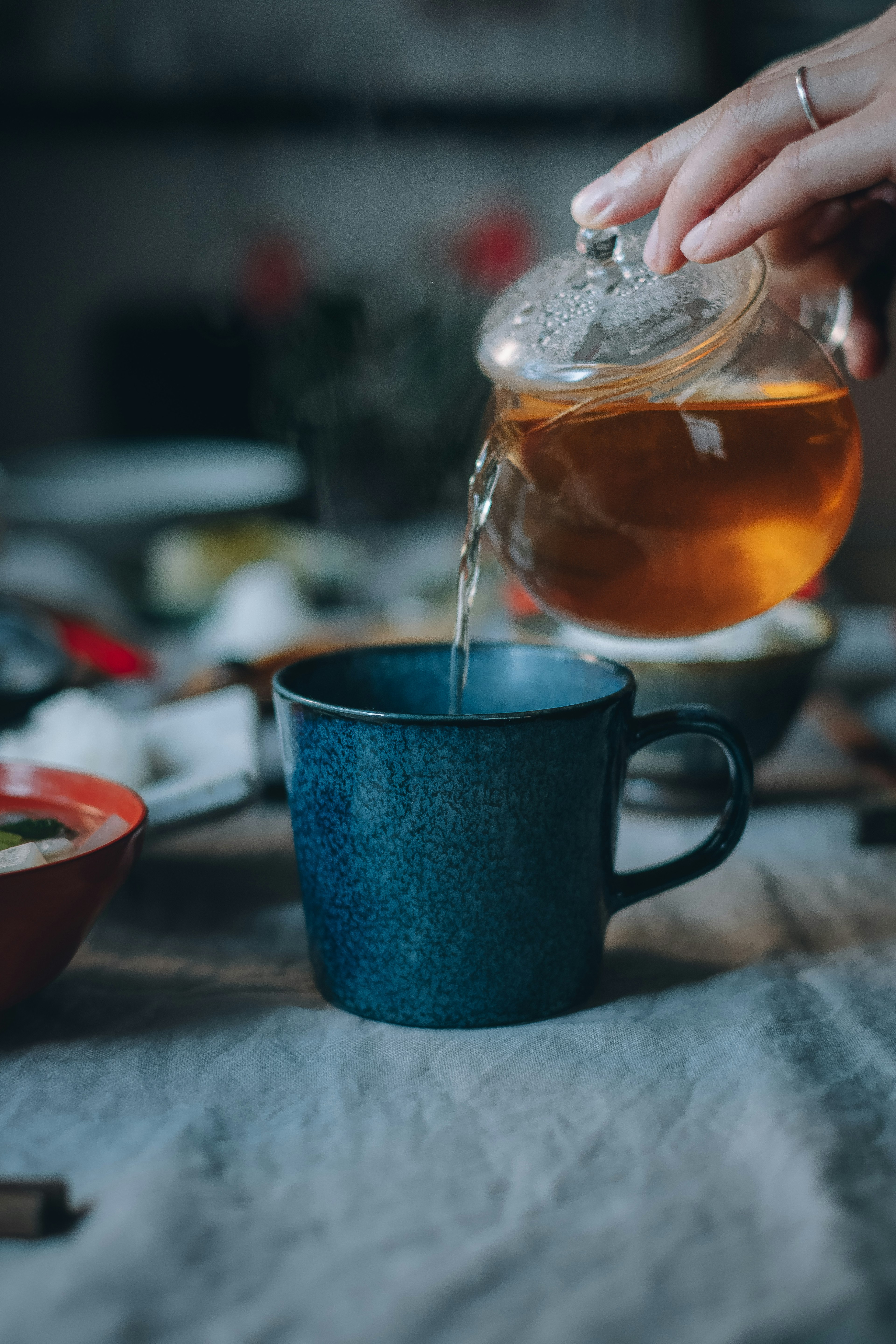 A hand pouring tea from a glass pot into a blue cup with surrounding dishes