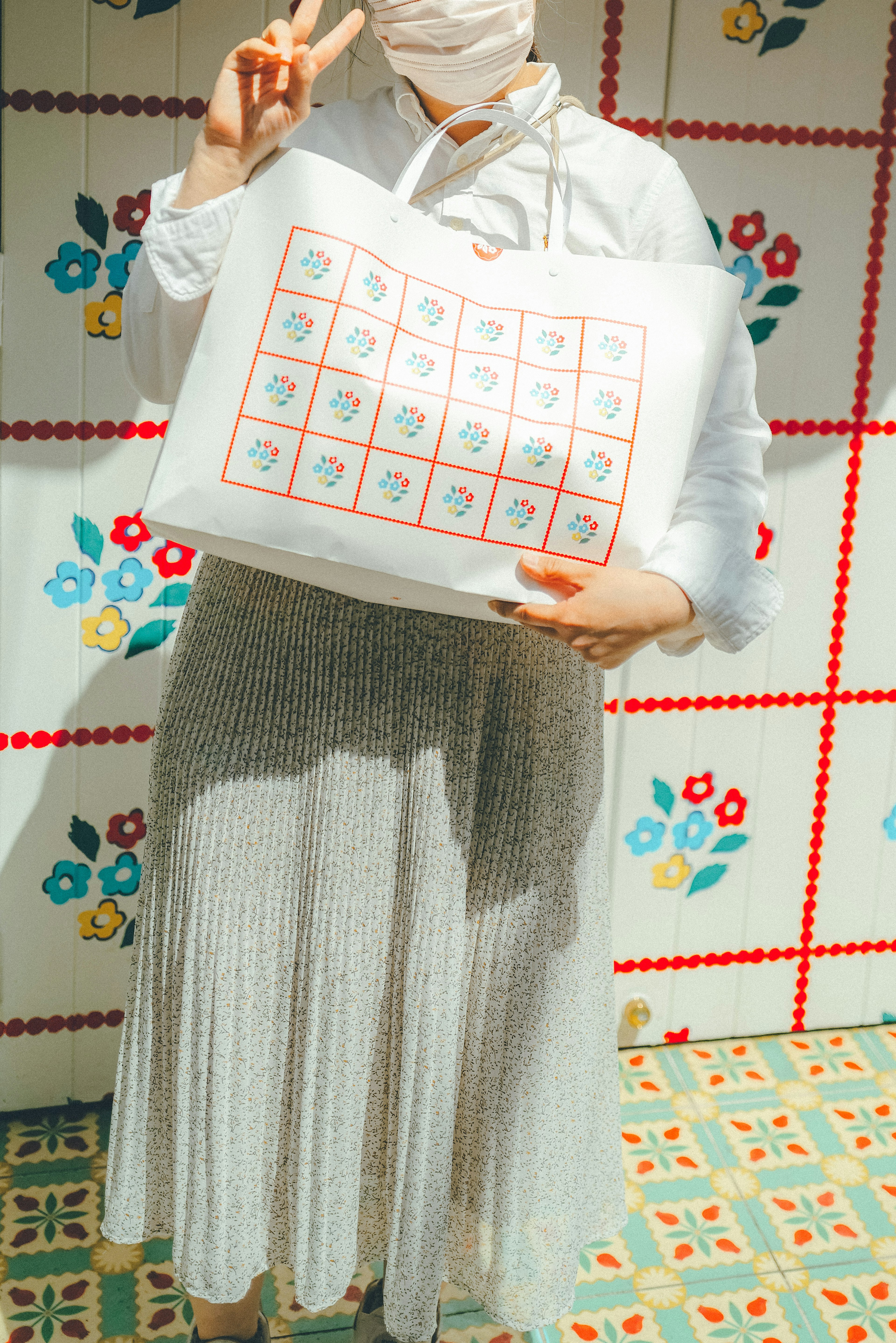 A woman in white clothing wearing a mask holding a floral patterned bag and making a peace sign