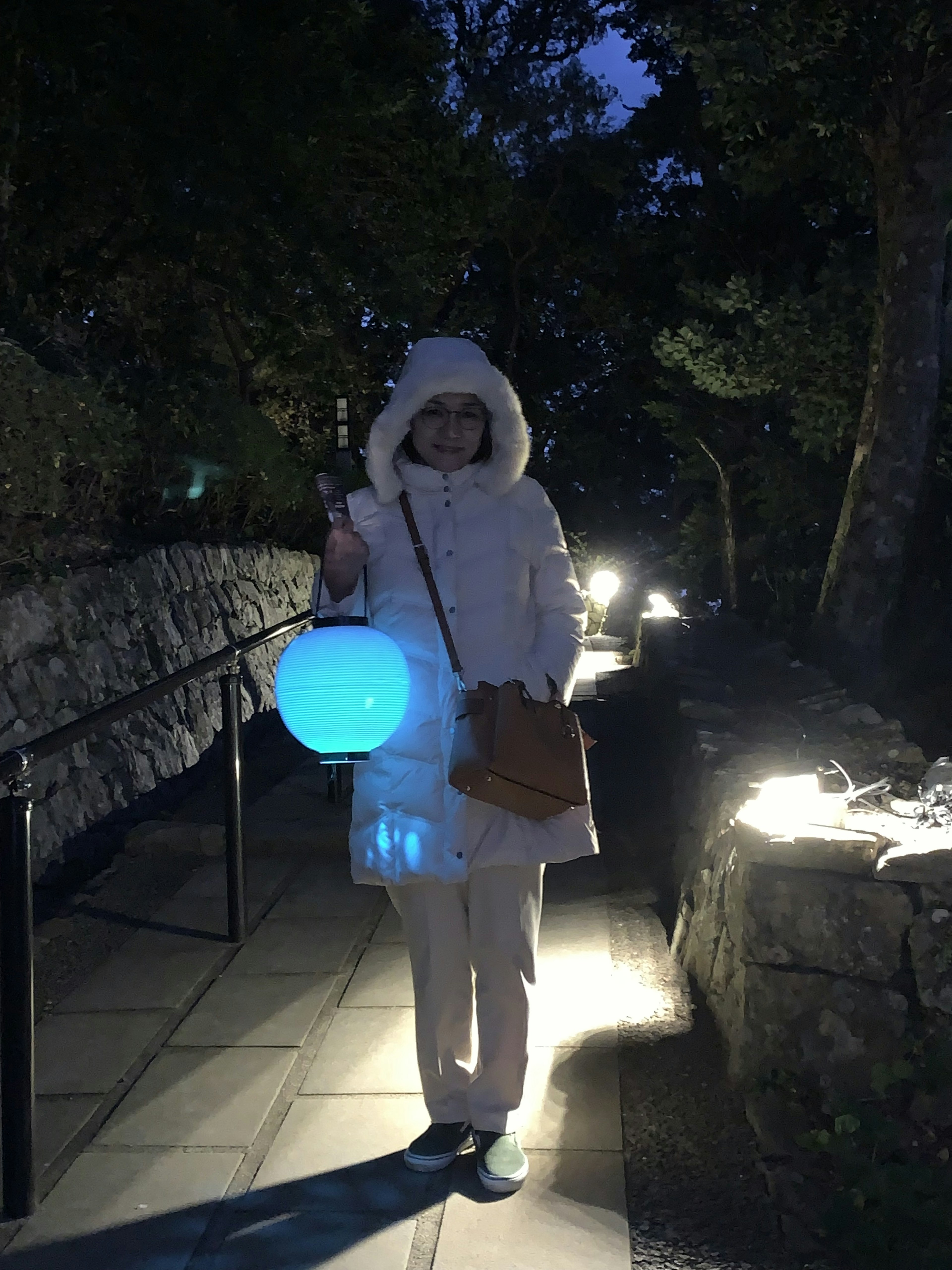 A woman in a white coat walking on a nighttime path holding a blue lantern