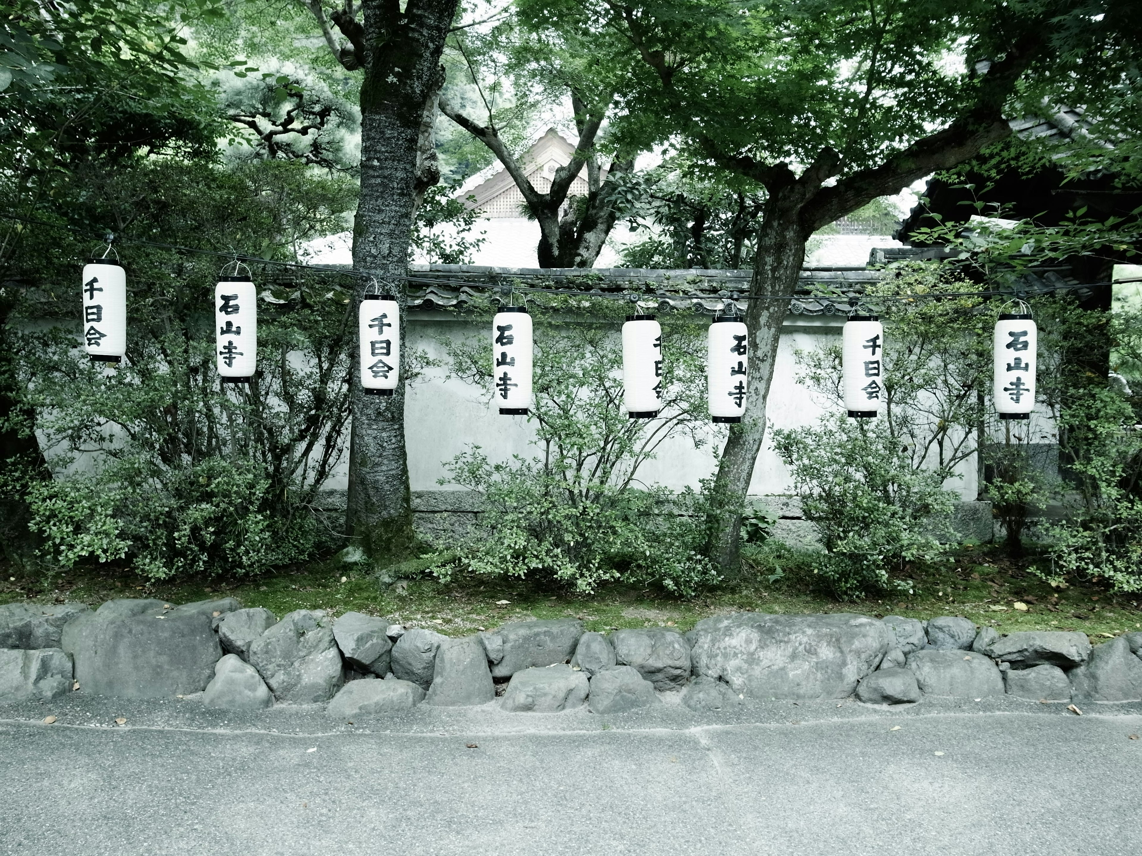 Japanische Schilder, die an einer weißen Wand hängen, umgeben von grünen Bäumen und einer Steinmauer