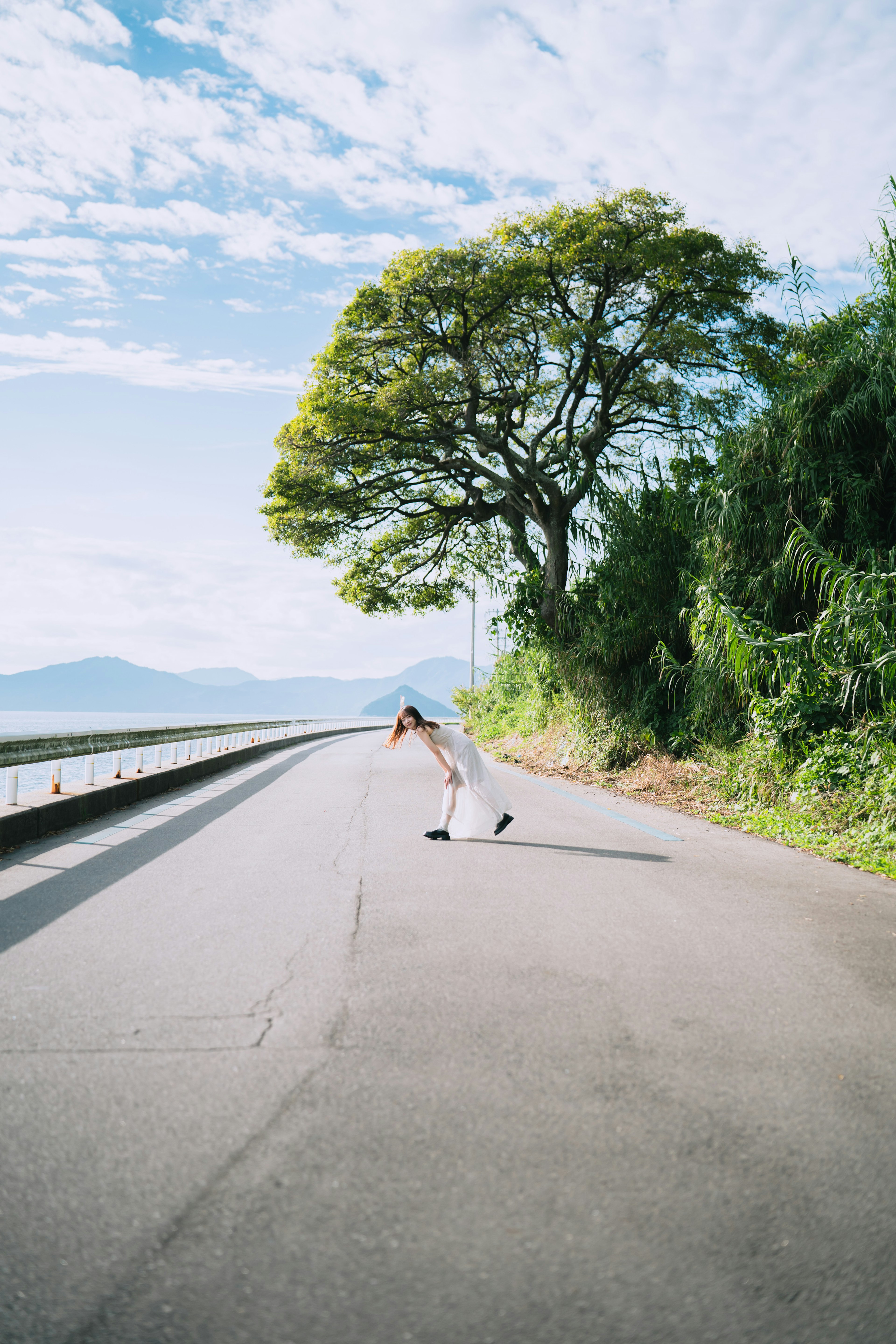 Mujer agachada en una carretera bajo un árbol verde y un cielo azul