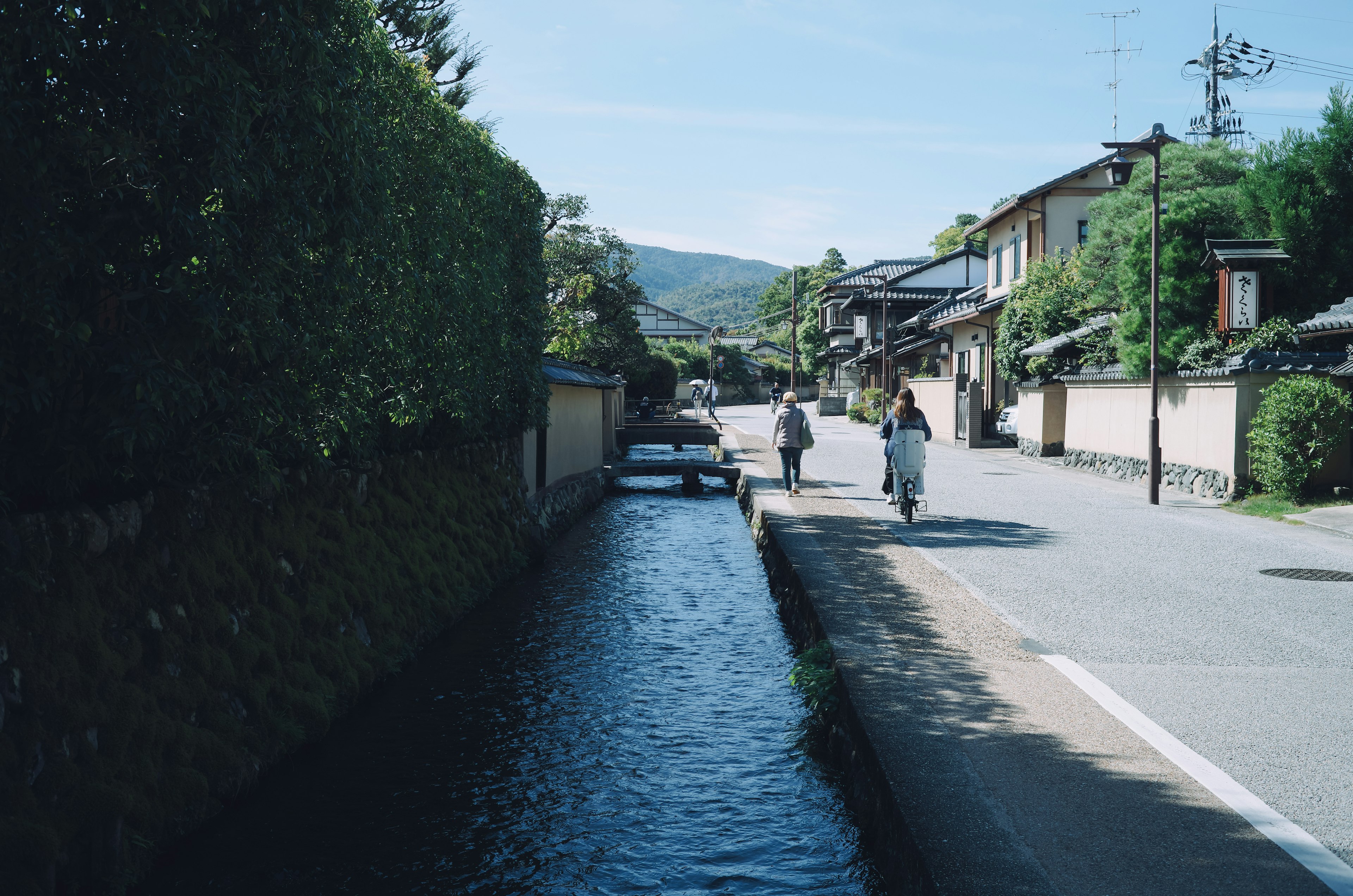 People walking along a quiet riverside path with lush greenery