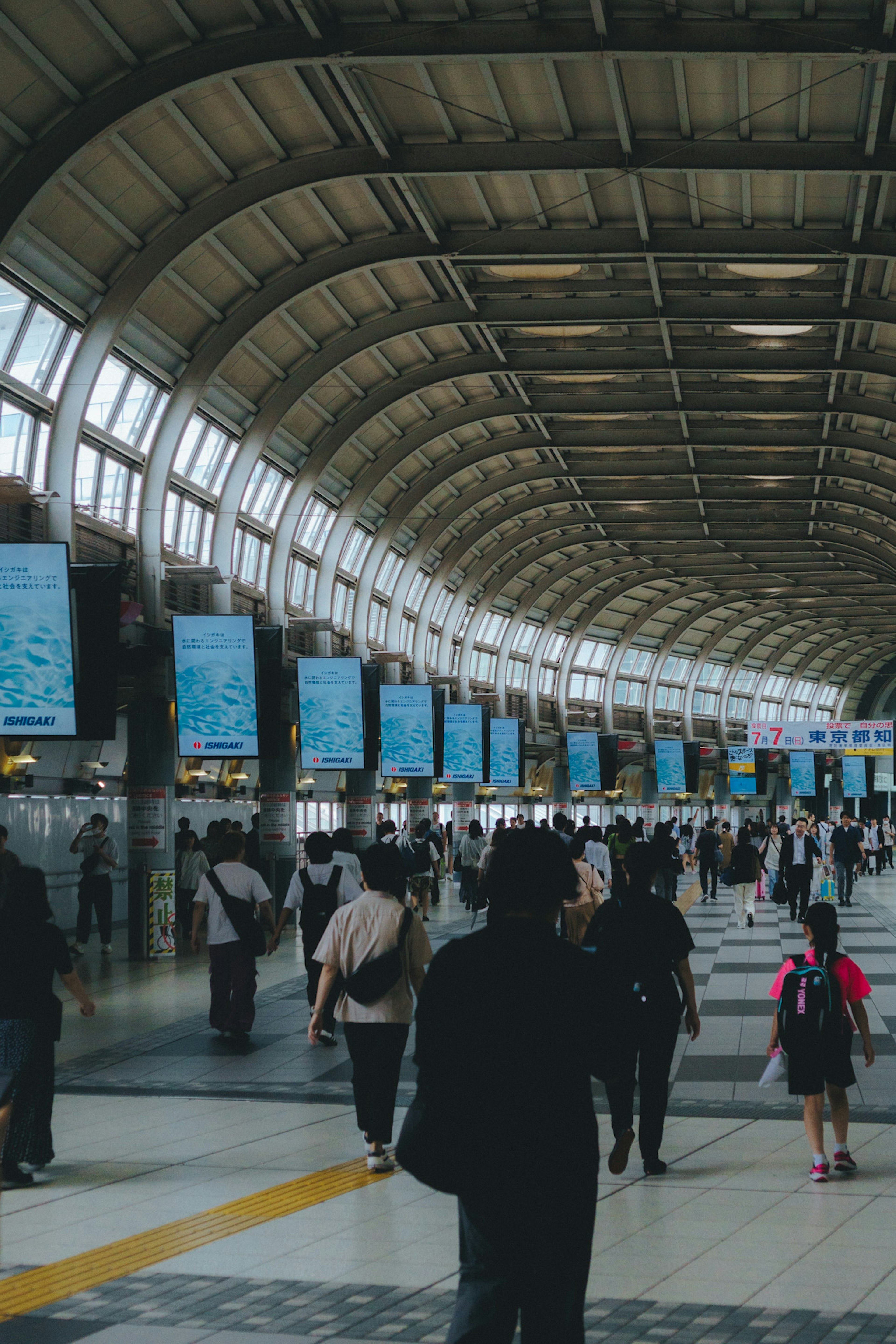 Estación de tren concurrida con personas caminando bajo un techo curvado y anuncios azules