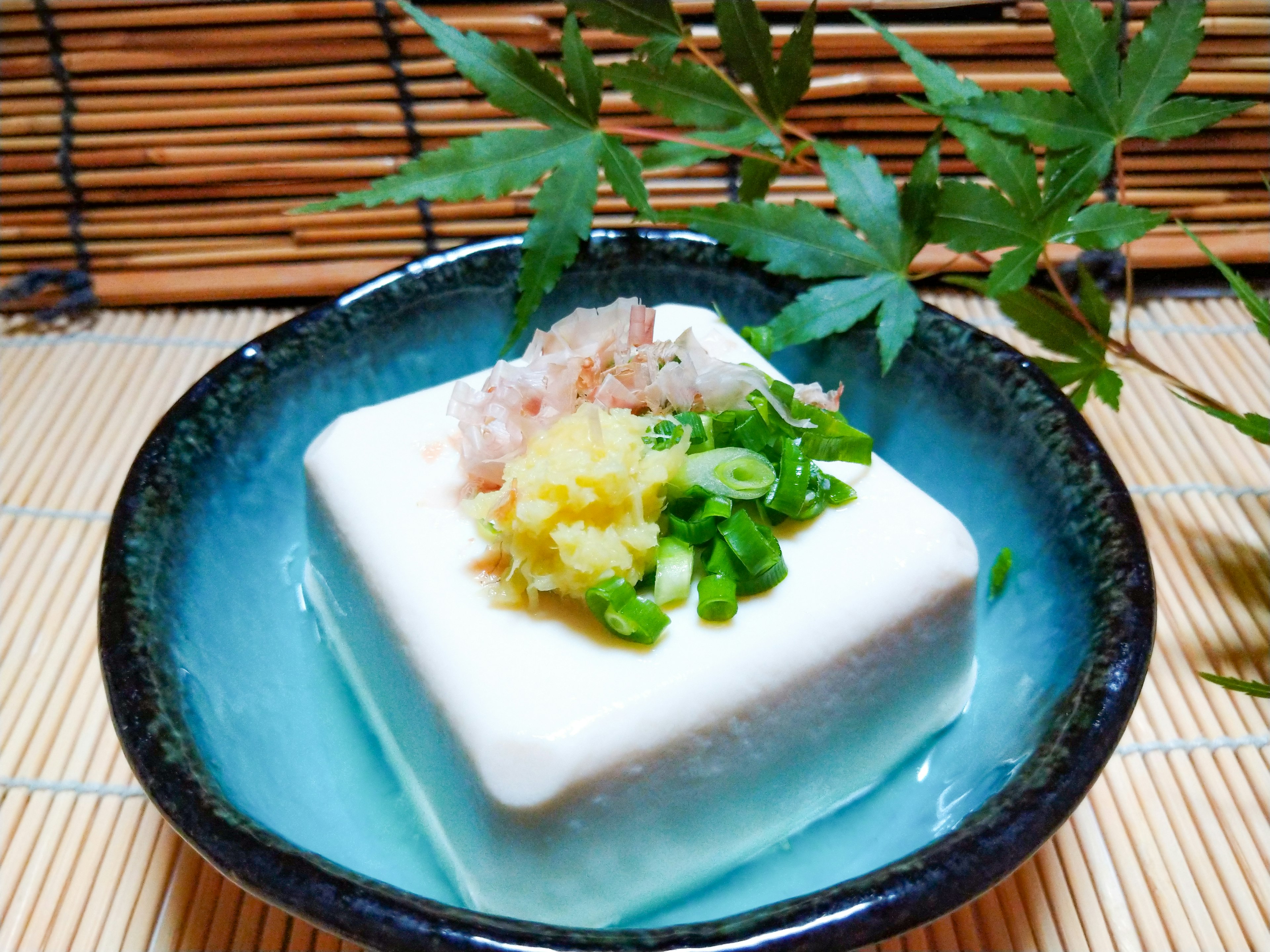 Tofu servi dans un plat bleu garni de gingembre, d'oignons verts et de flocons de bonito