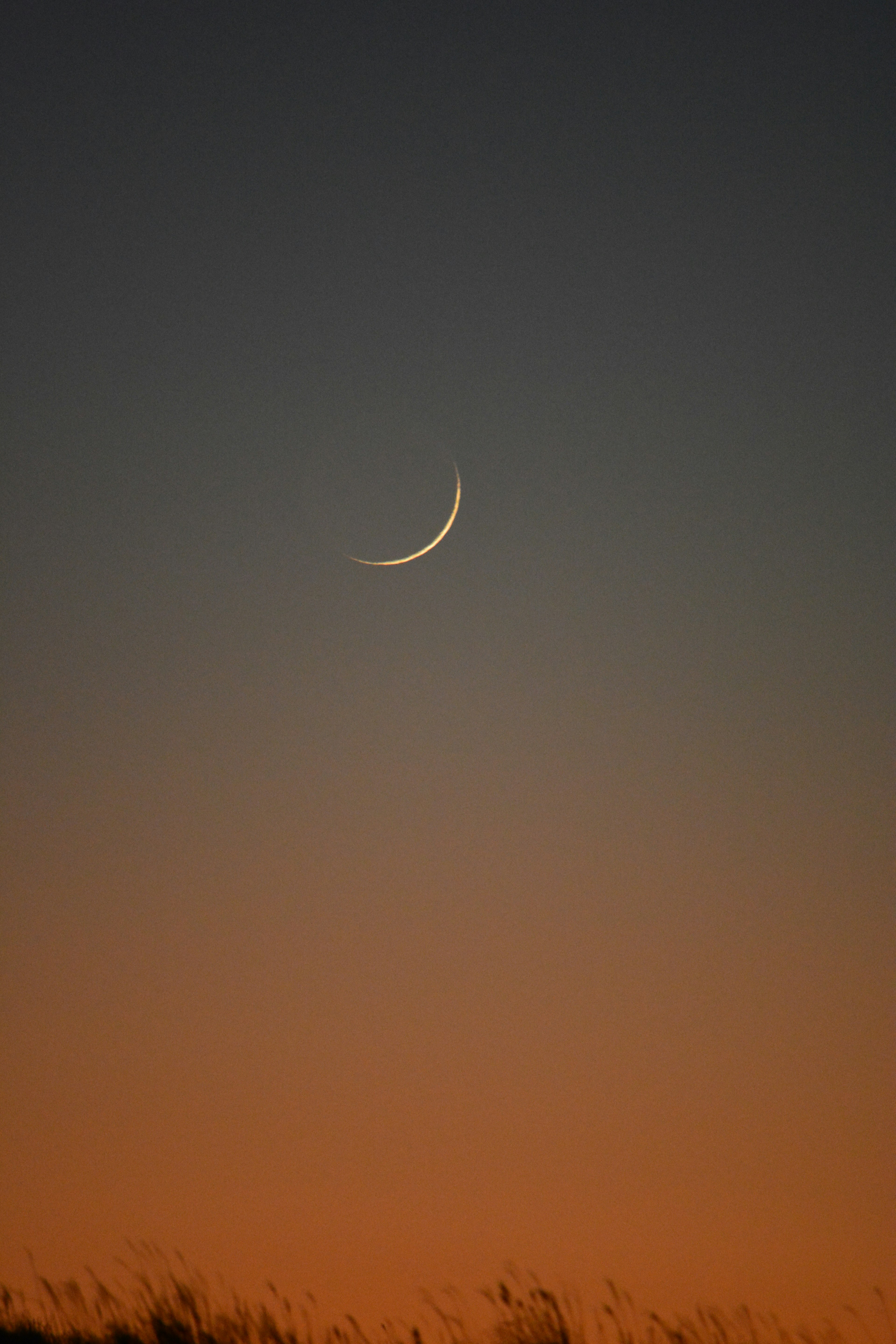 Bulan sabit tipis di langit senja dengan nuansa oranye