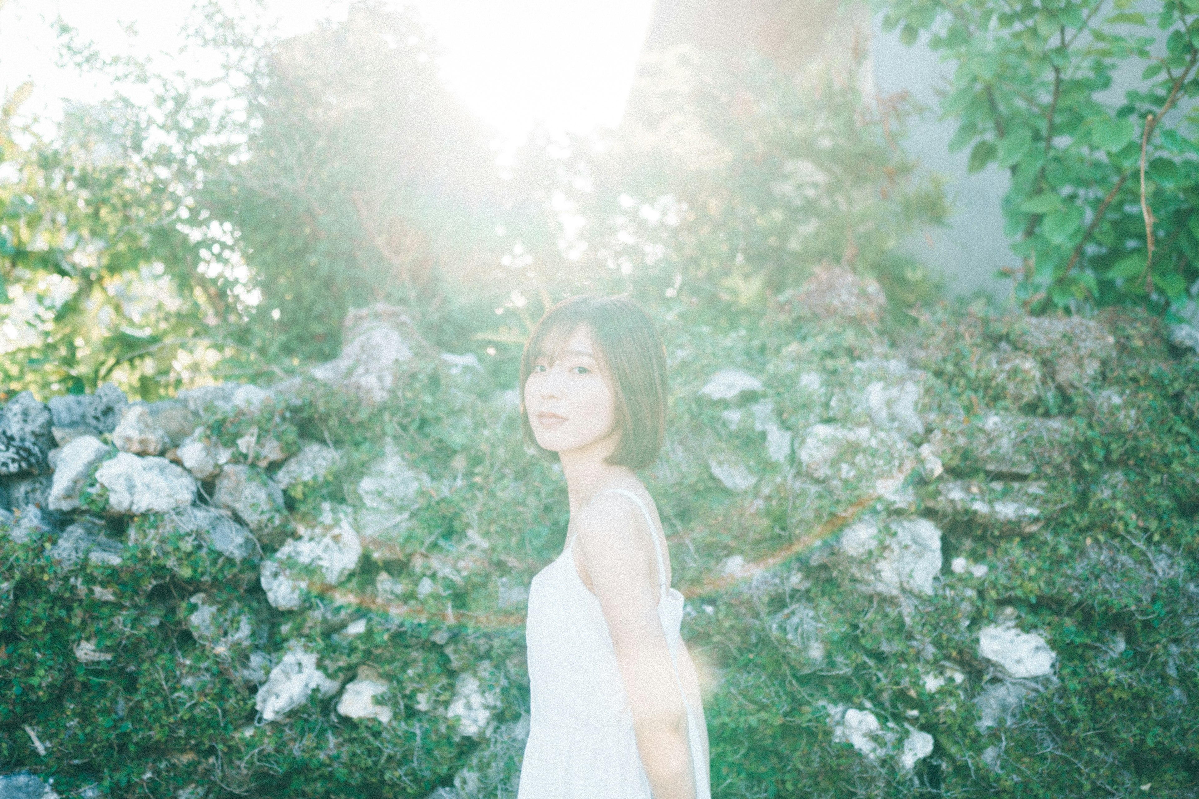 A woman in a white dress stands against a sunlit green backdrop