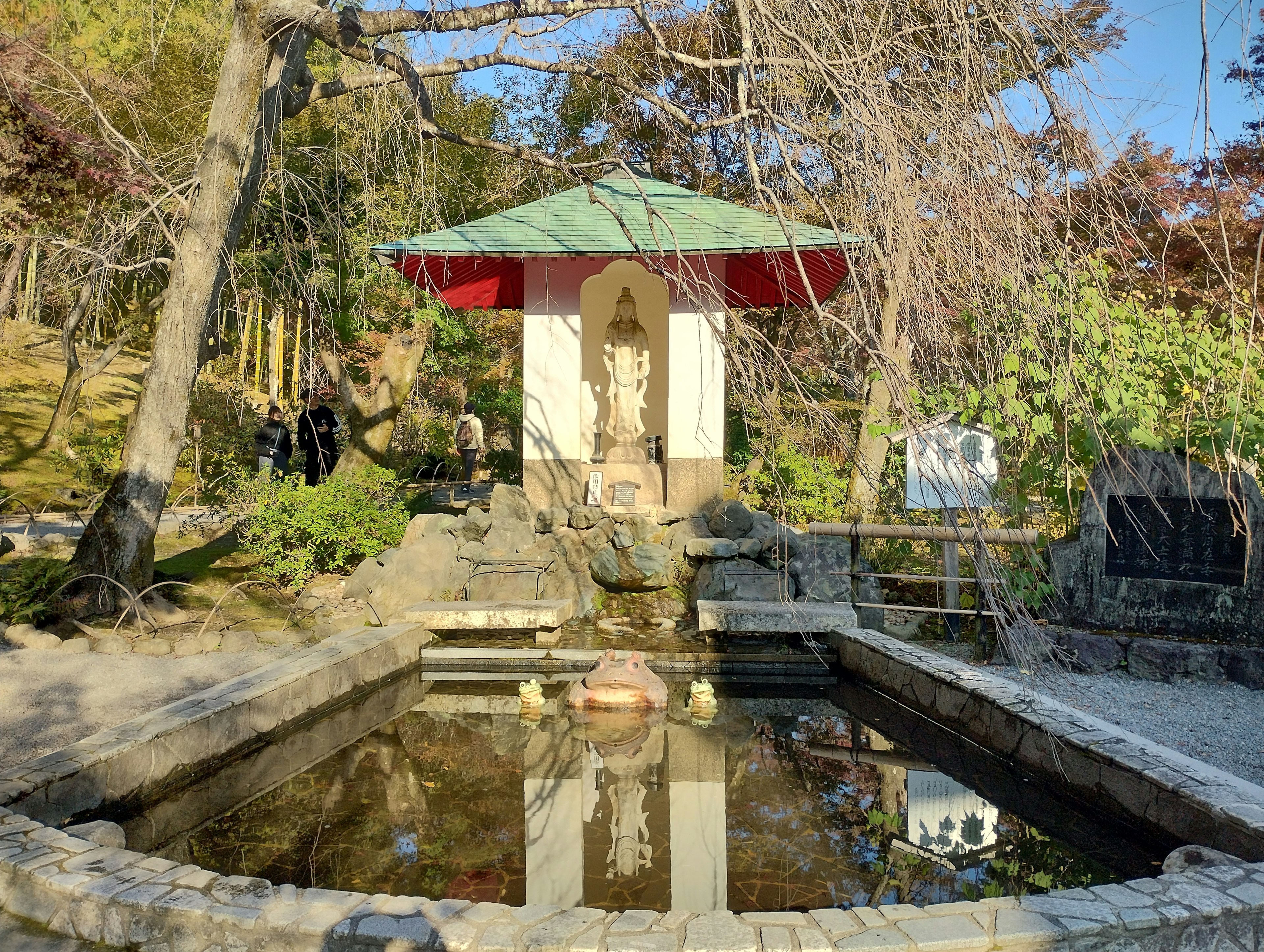 Pemandangan taman dengan paviliun kecil atap hijau dan patung yang terpantul di air