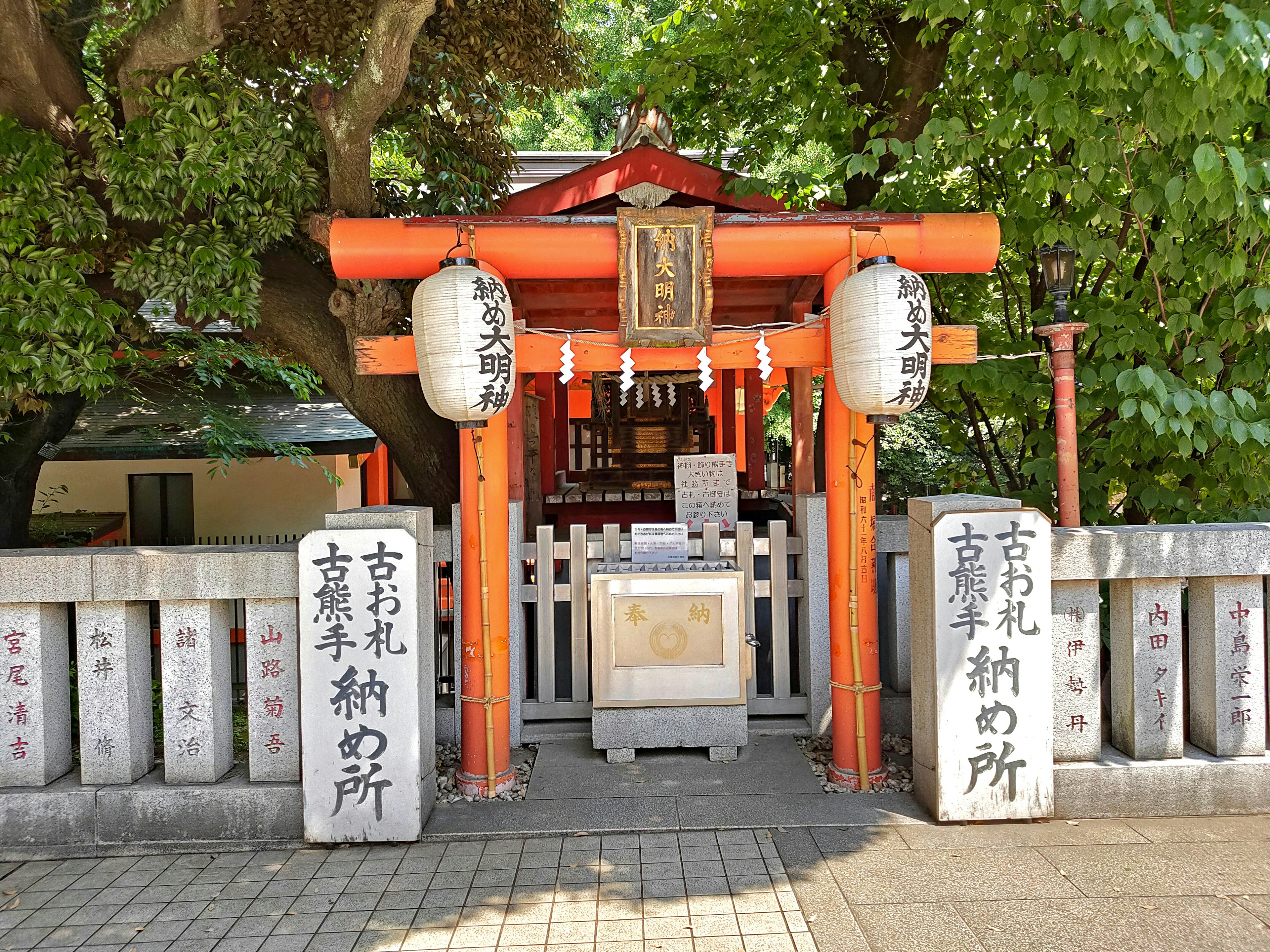 Eingang eines Schreins mit rotem Torii und Laternen, umgeben von grünen Bäumen