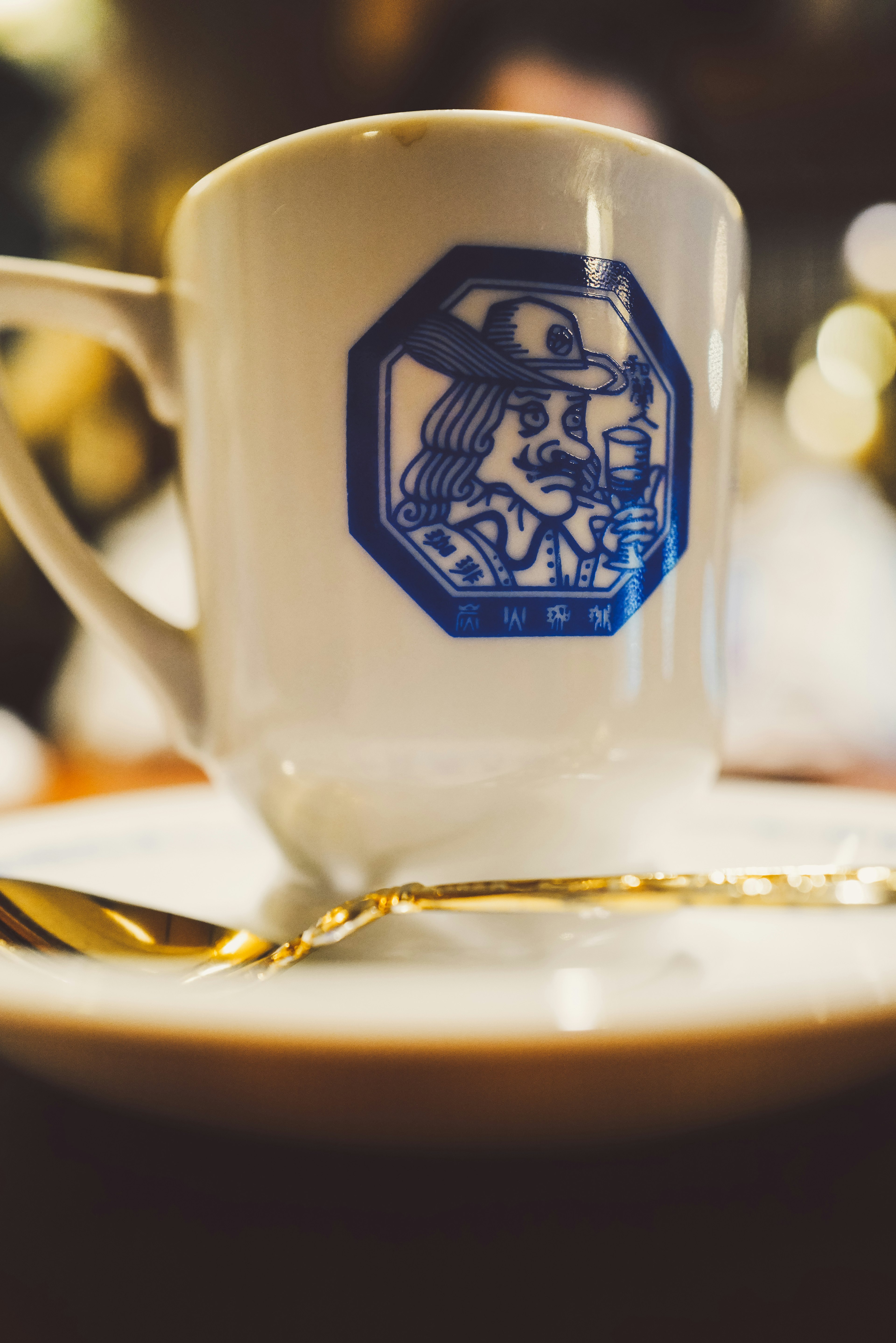 Taza de café blanca con diseño azul y cuchara dorada en un plato