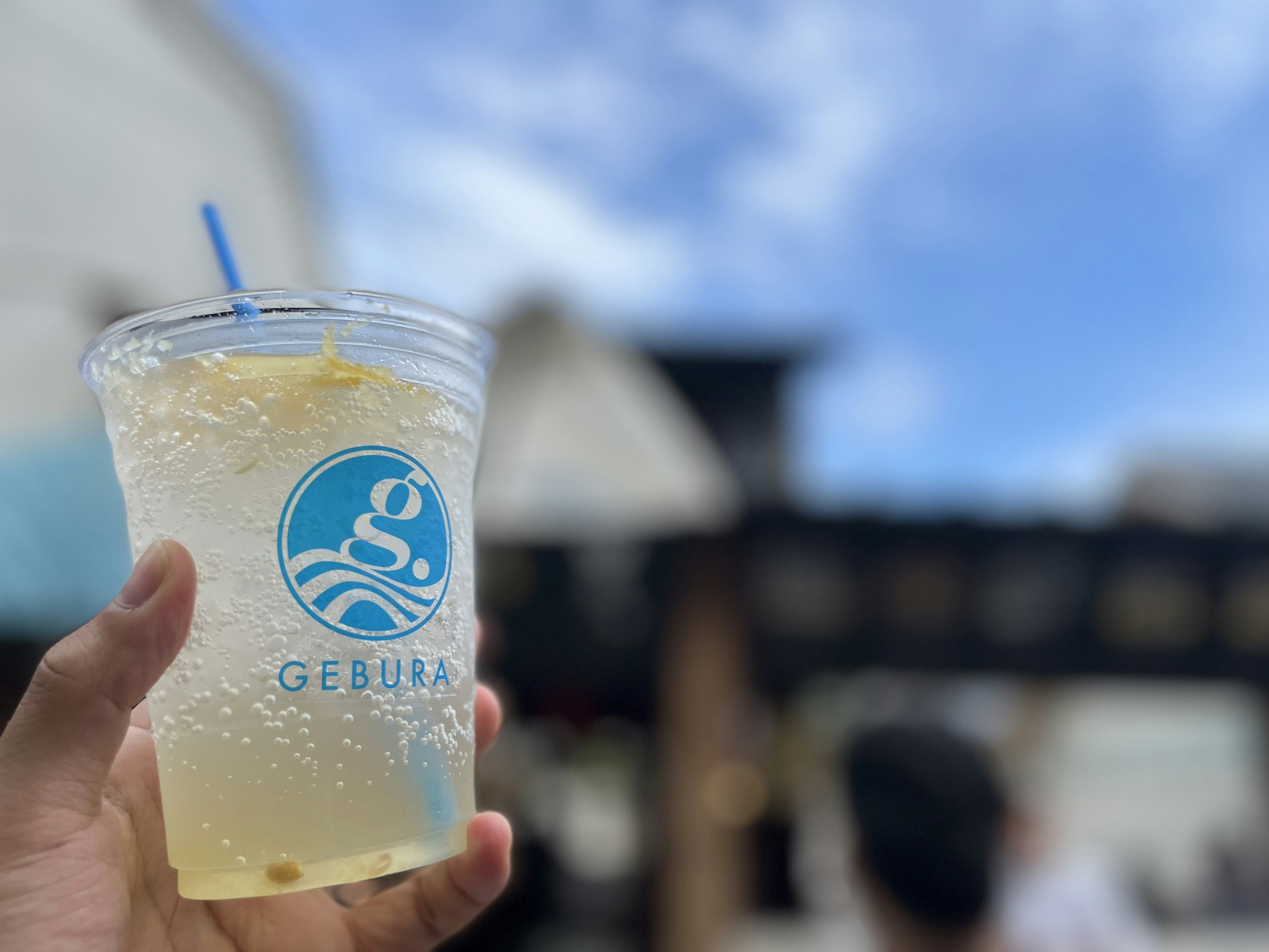 A hand holding a clear beverage cup with a logo and blue sky in the background