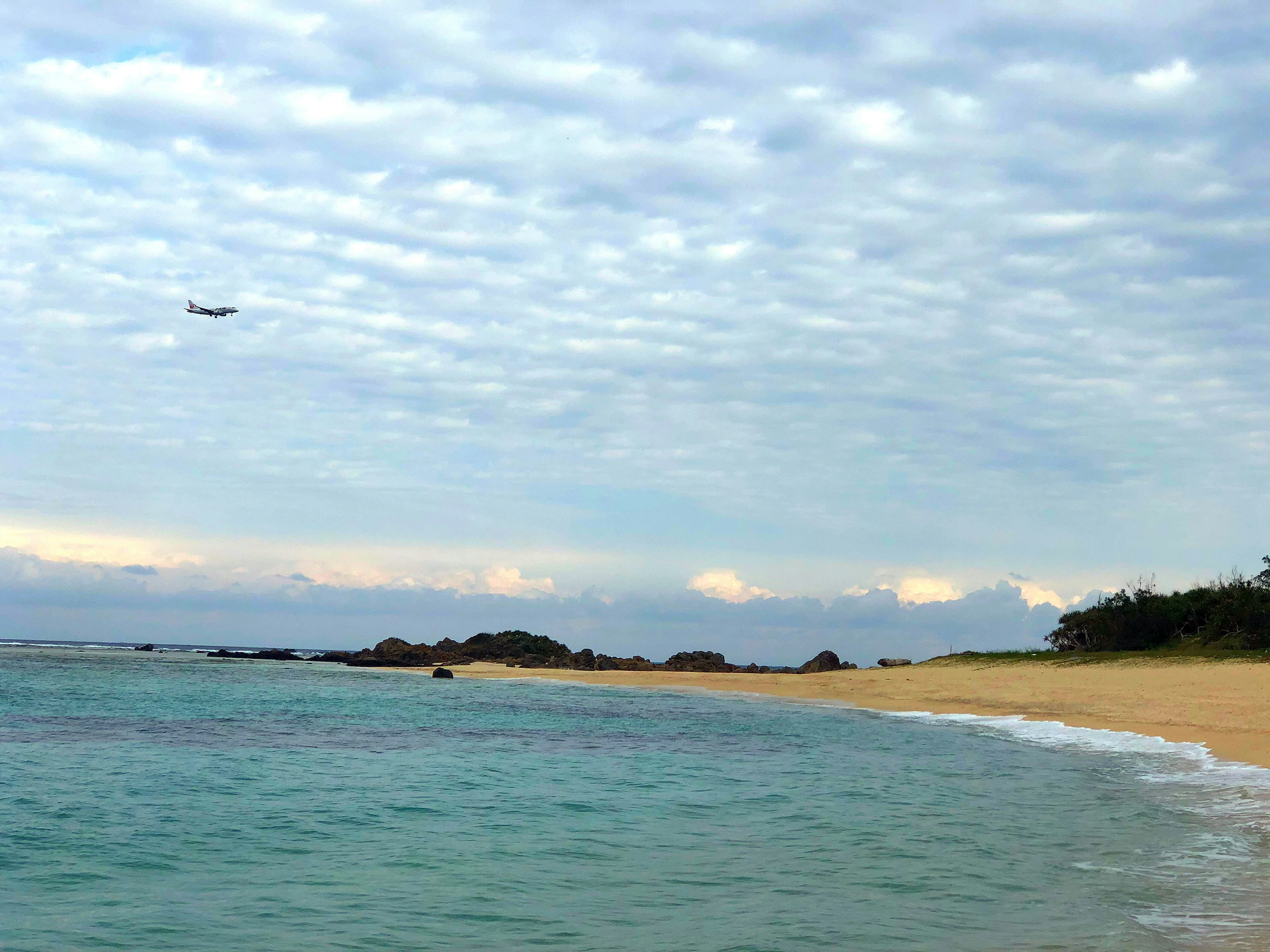 Szenischer Strandblick mit türkisfarbenem Wasser und bewölktem Himmel Flugzeug in der Ferne