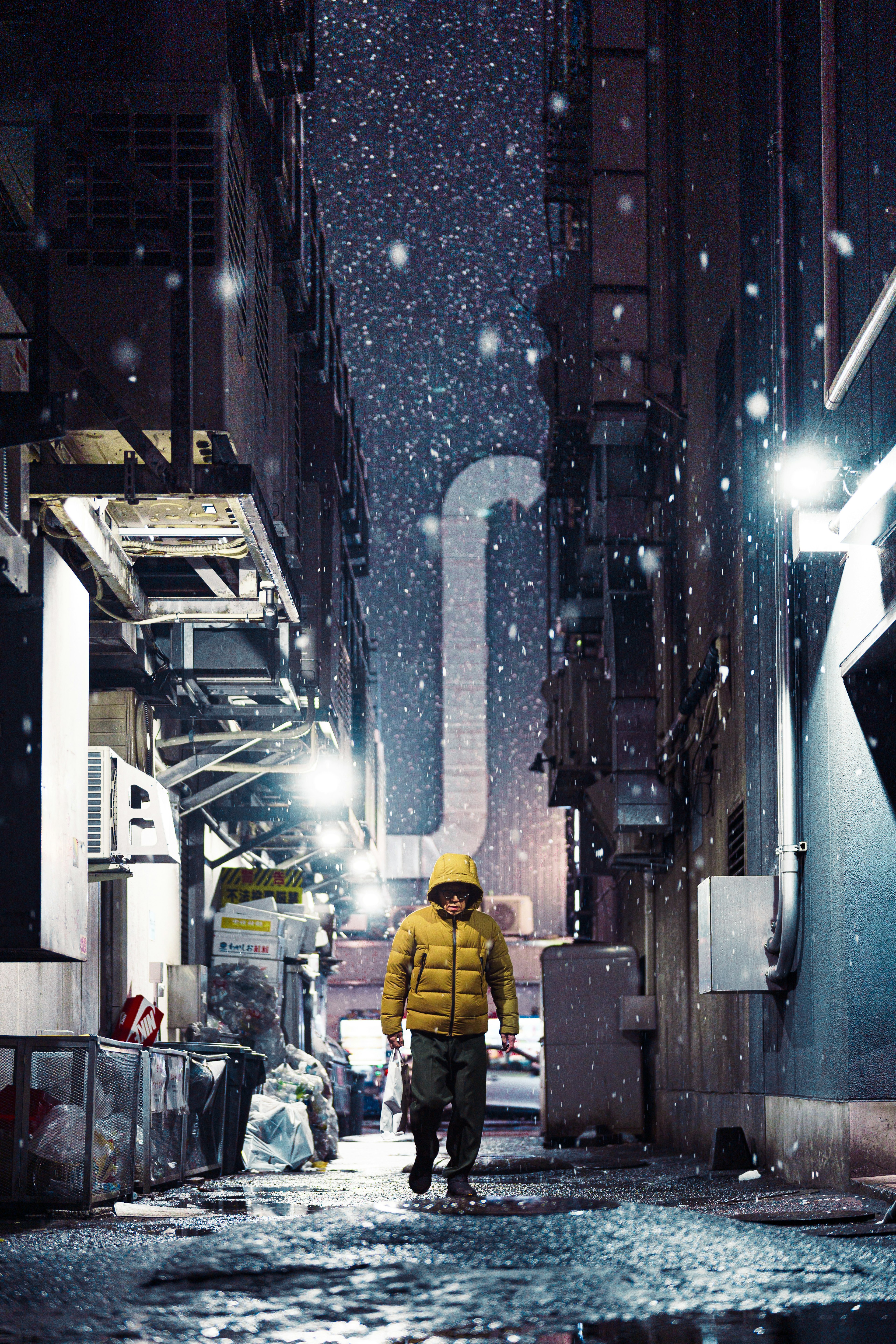 Person in yellow jacket walking in narrow alley during snowfall at night