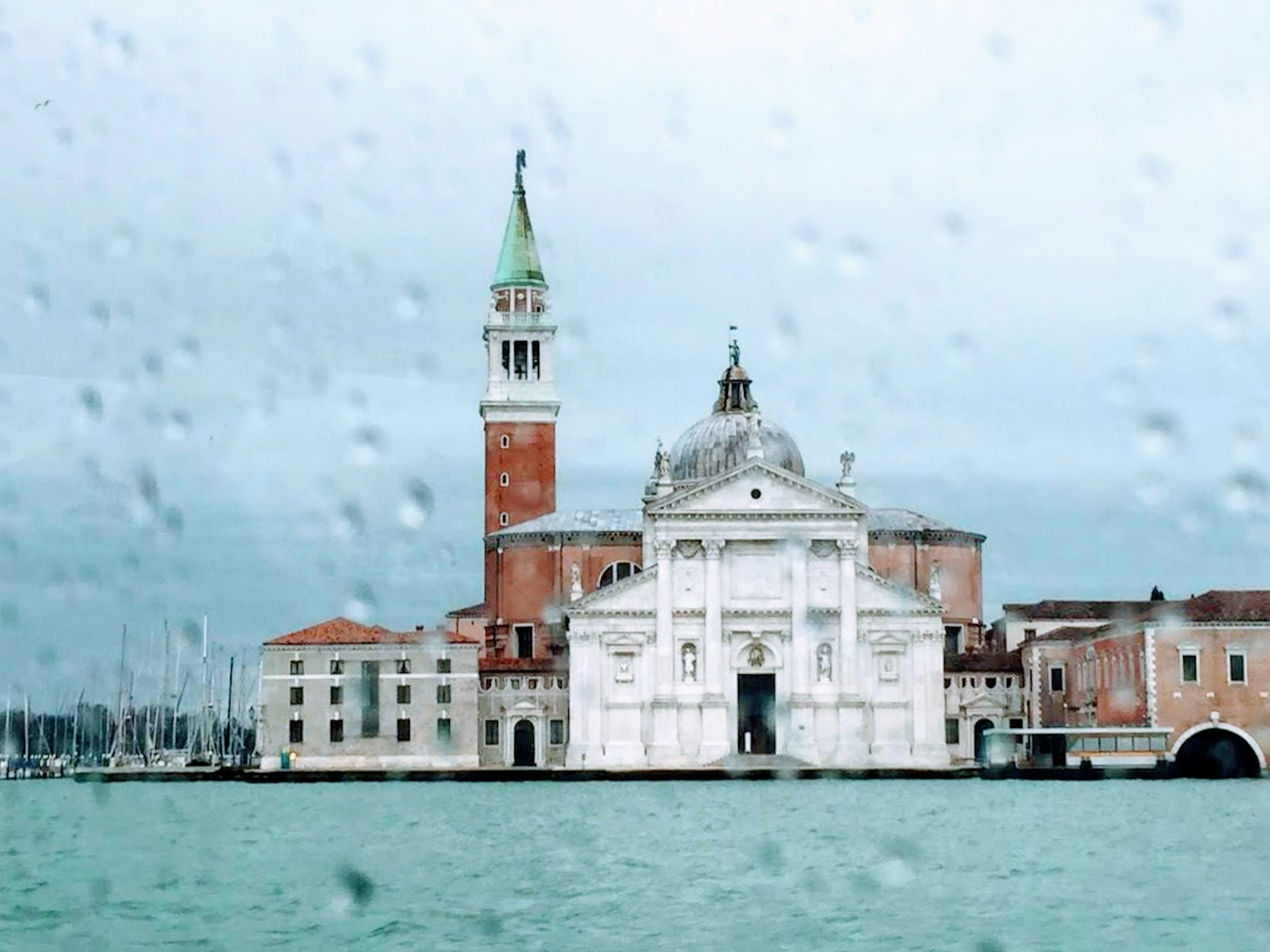 Vue d'un bâtiment blanc et d'un clocher à Venise à travers des gouttes de pluie sur une fenêtre