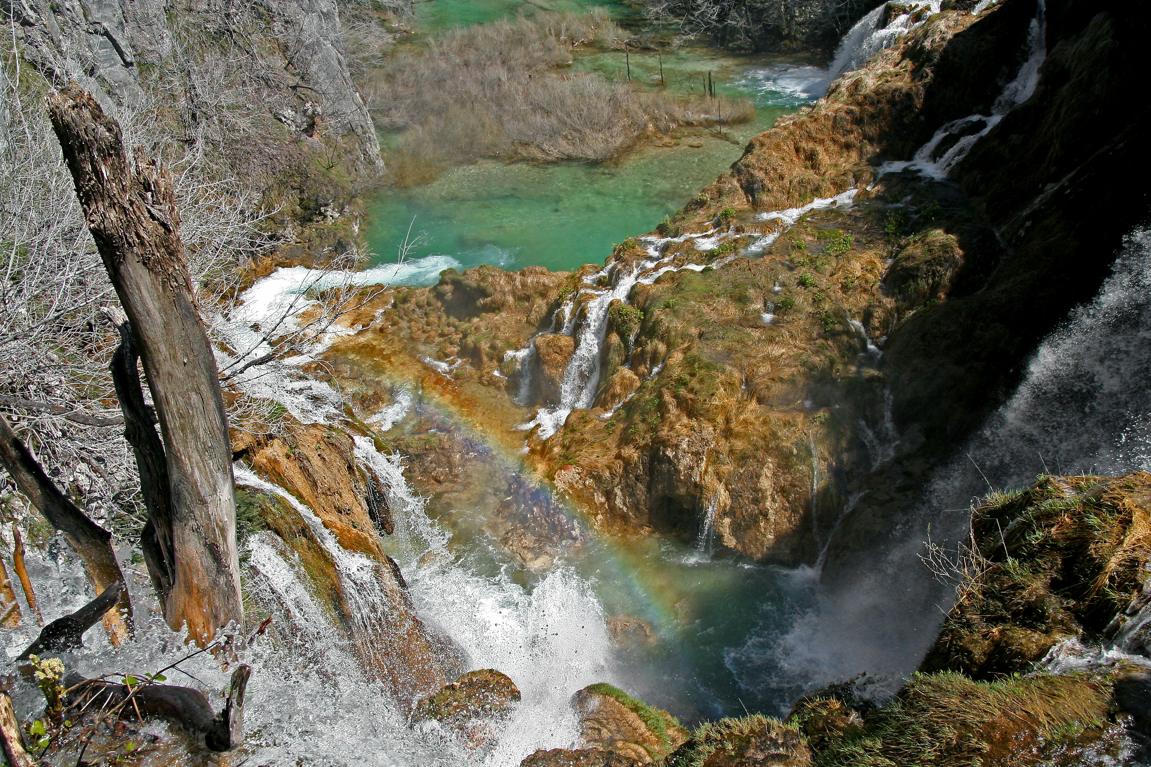 Blick von oben auf einen Wasserfall mit grünem Wasser und einem Regenbogen zwischen den Felsen