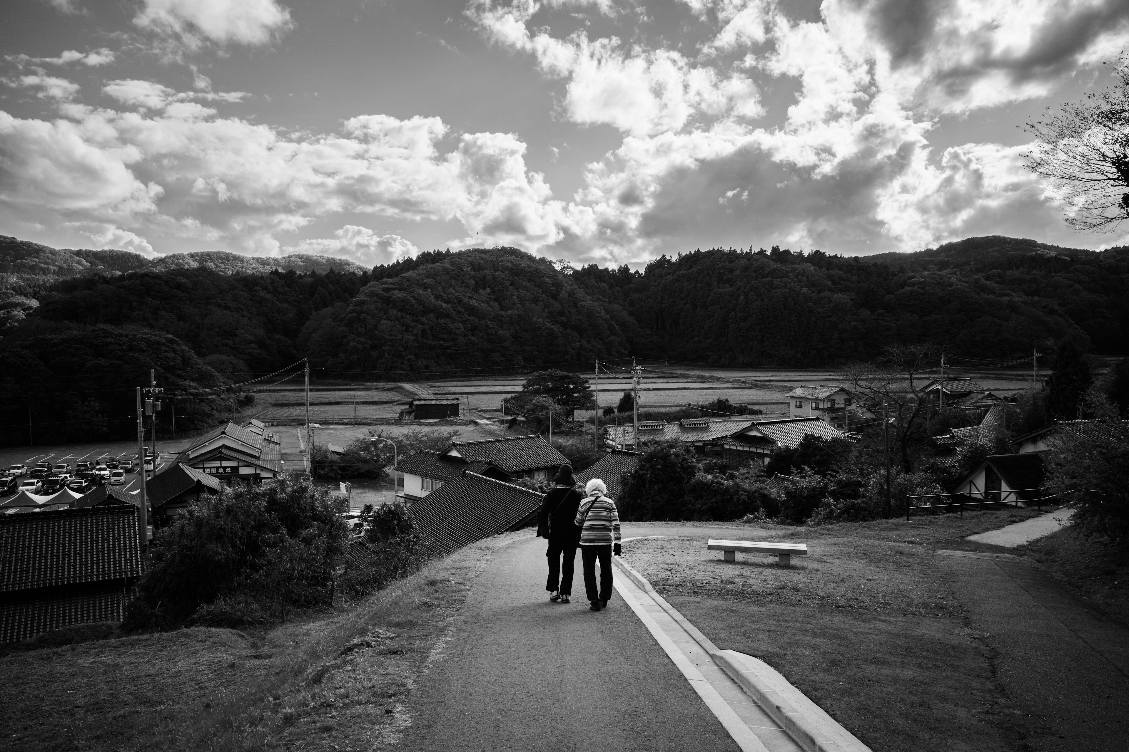 Deux personnes marchant le long d'un chemin rural avec des montagnes et des nuages en arrière-plan en noir et blanc