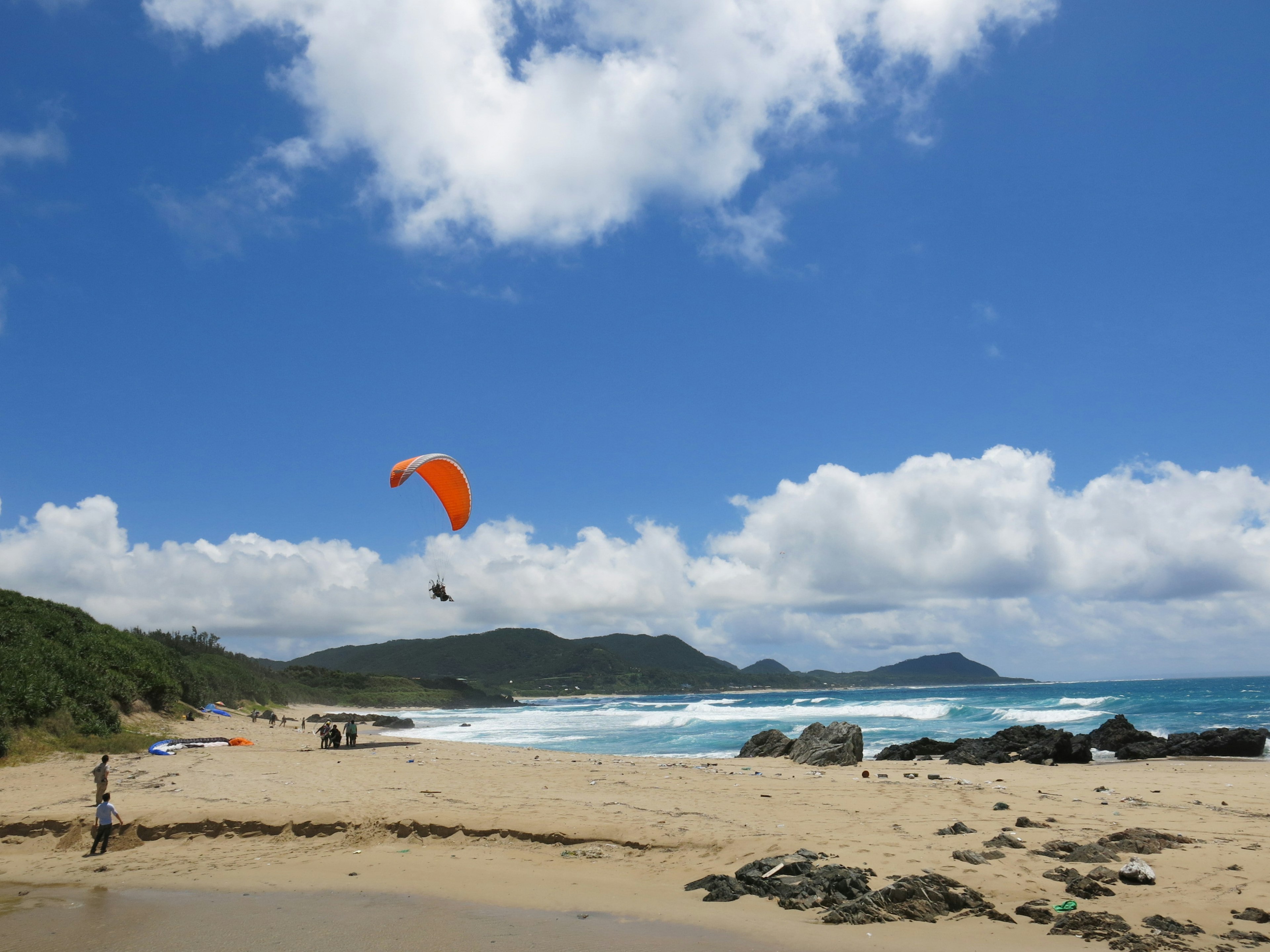 Strandszene mit einem orangefarbenen Paraglider, der unter einem blauen Himmel und weißen Wolken fliegt