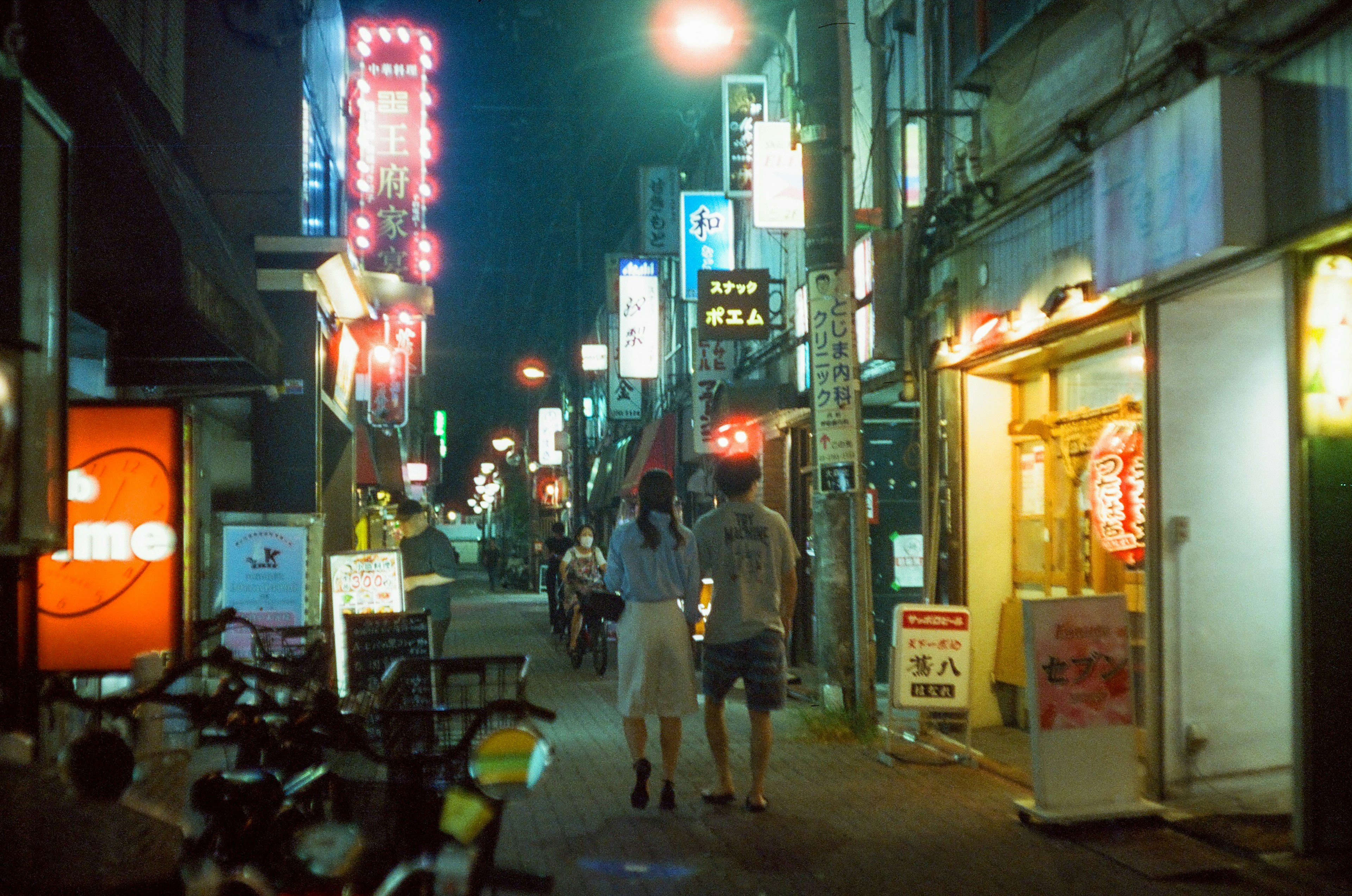 夜の街並みを歩くカップルの写真 ネオンの看板が輝く狭い通り