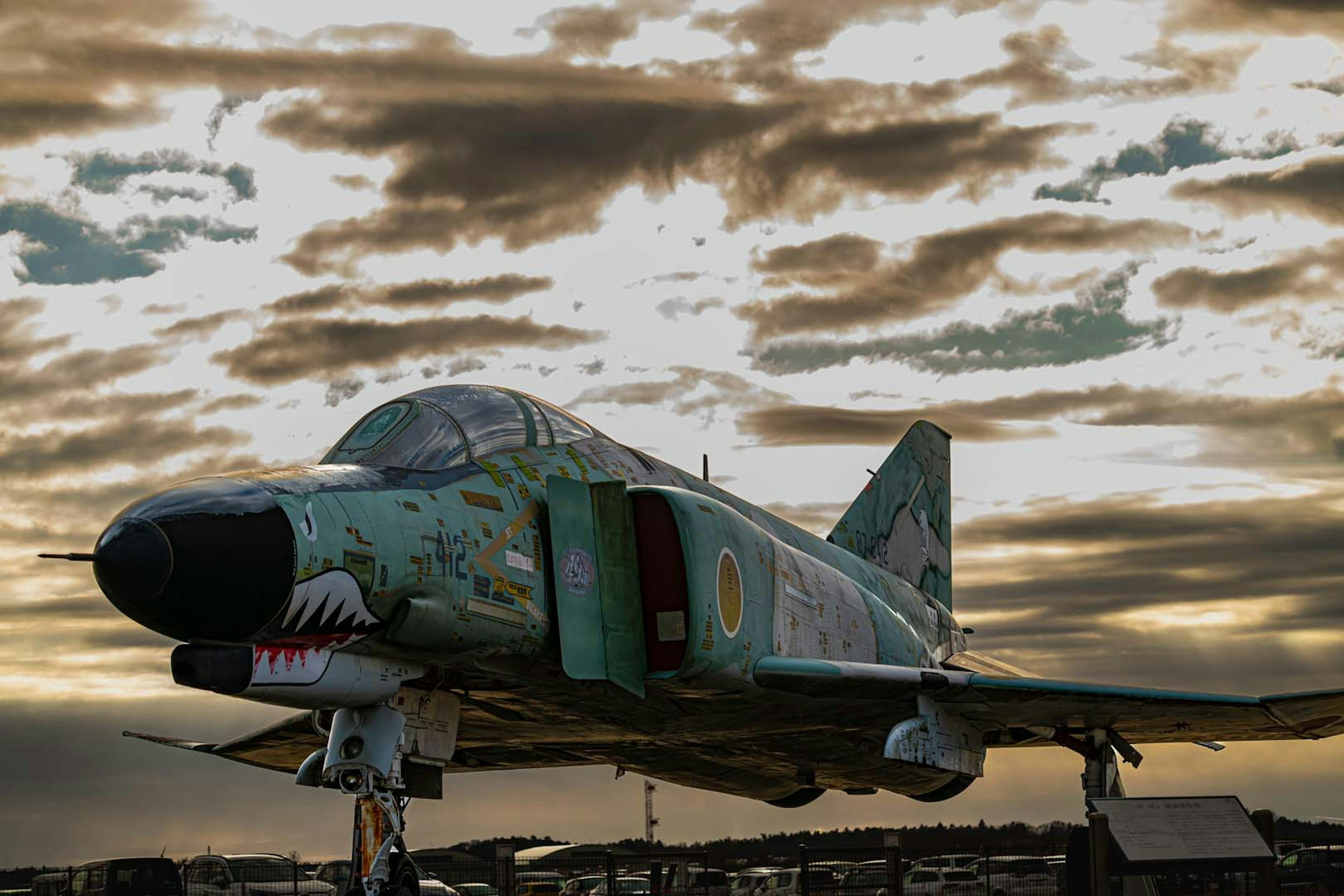 A fighter jet on display under a twilight sky featuring green camouflage and shark mouth design