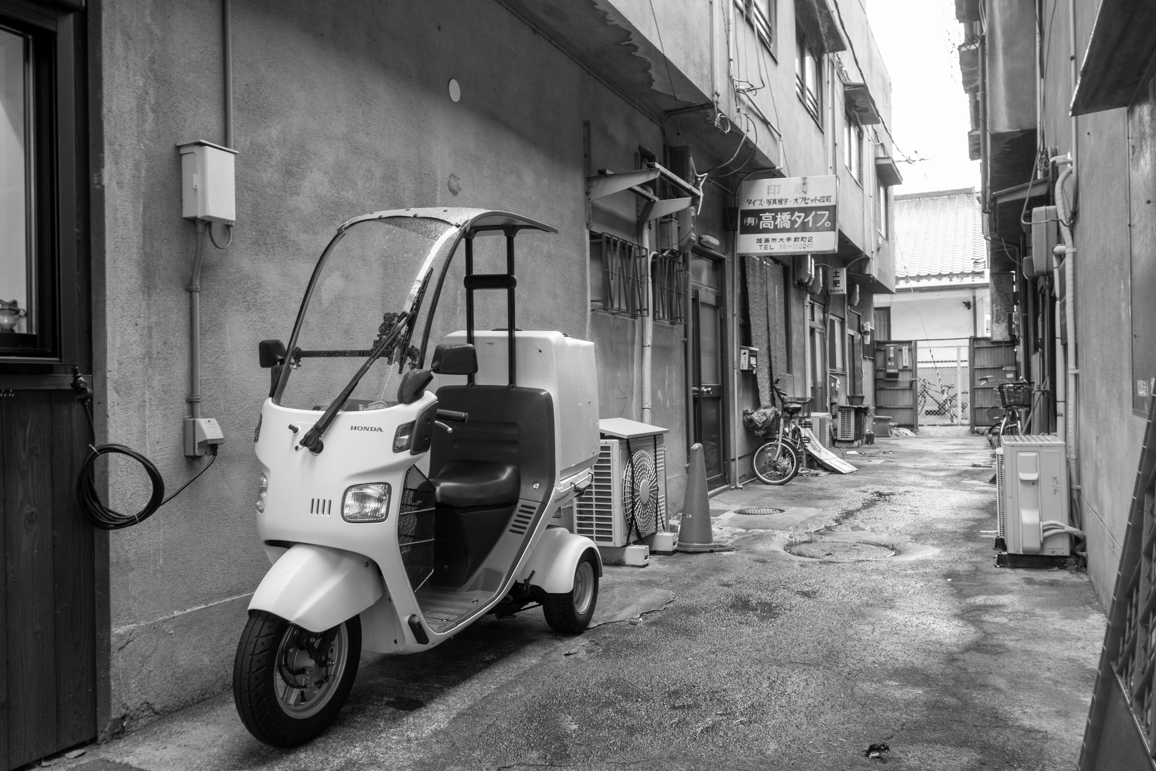 Gang sempit dengan kendaraan tiga roda putih yang diparkir dan bangunan tua