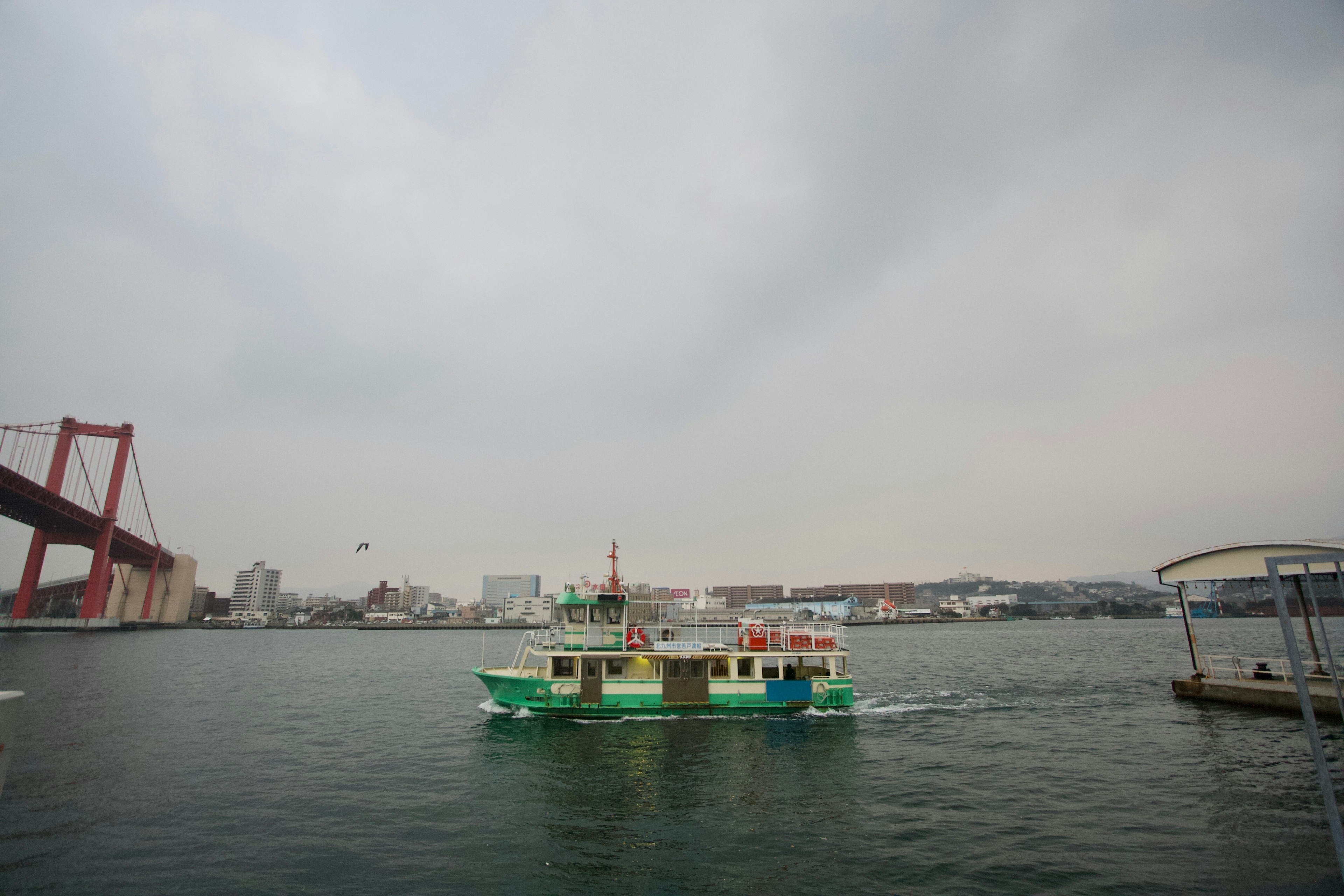 Grünes Fährschiff, das im Wasser nahe einem Hafen mit einer roten Brücke fährt