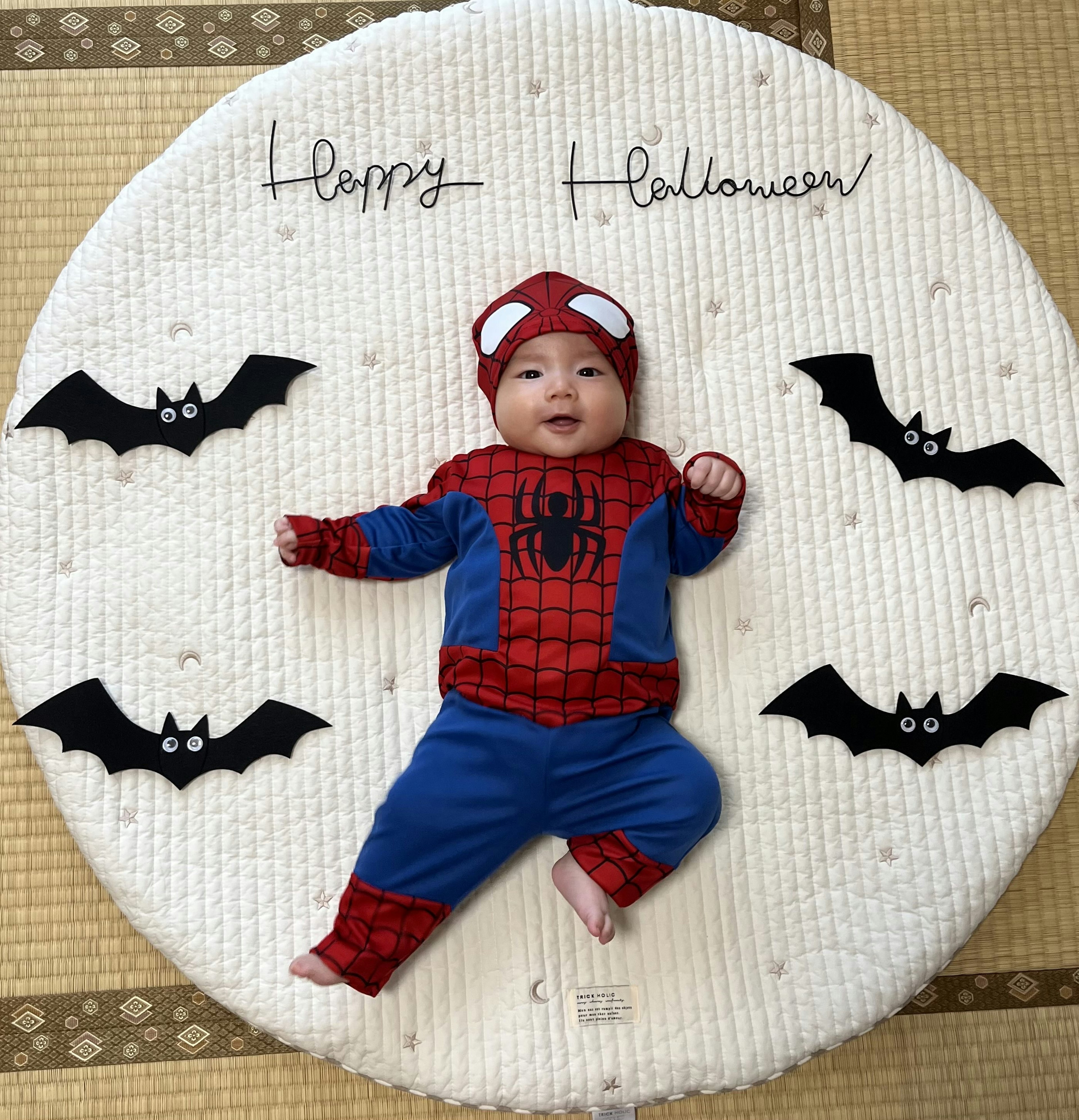 Un bebé vestido de Spider-Man acostado en una alfombra de Halloween rodeado de murciélagos