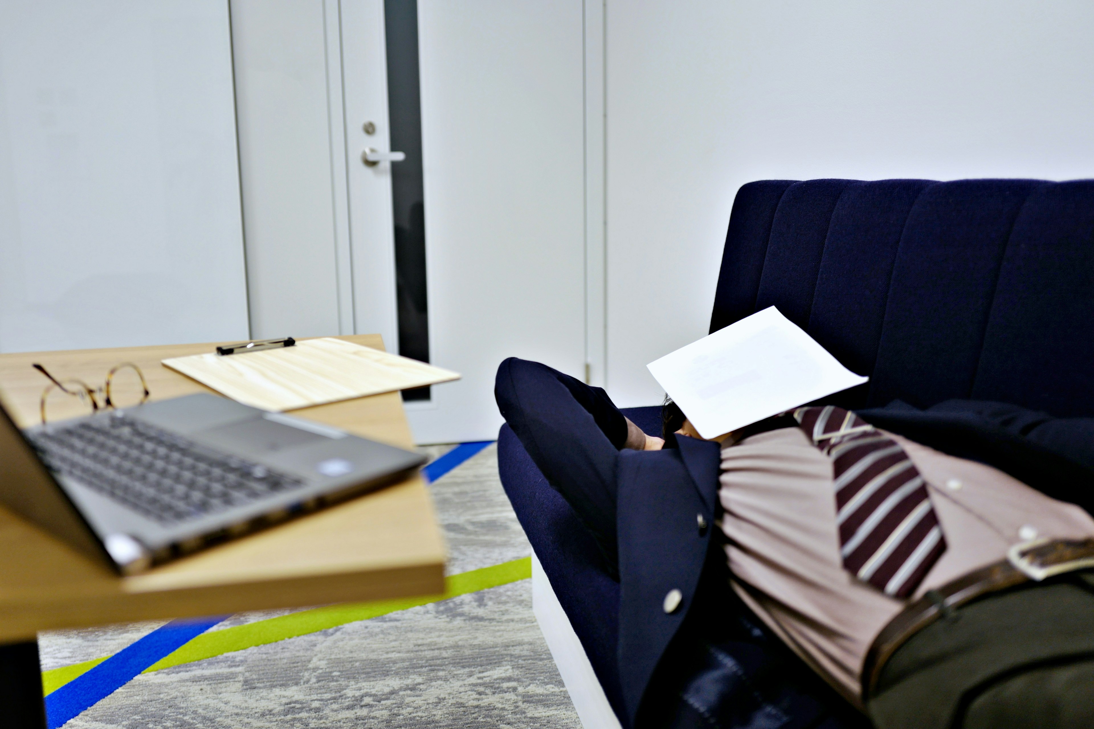 A man lying on a blue sofa with a laptop and documents on a desk