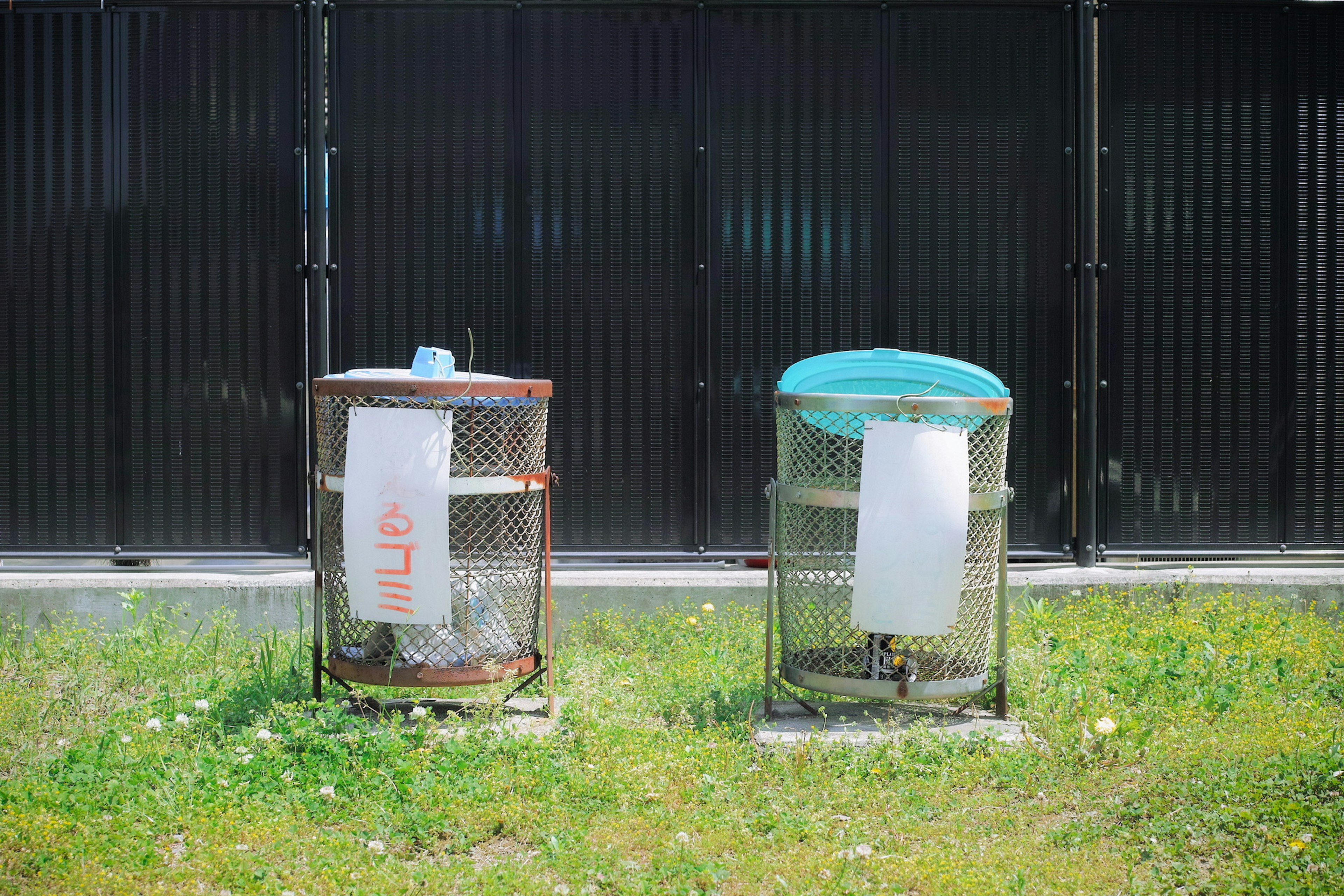 Deux poubelles sur de l'herbe l'une est marron et l'autre a un couvercle bleu l'arrière-plan présente une clôture en métal noir