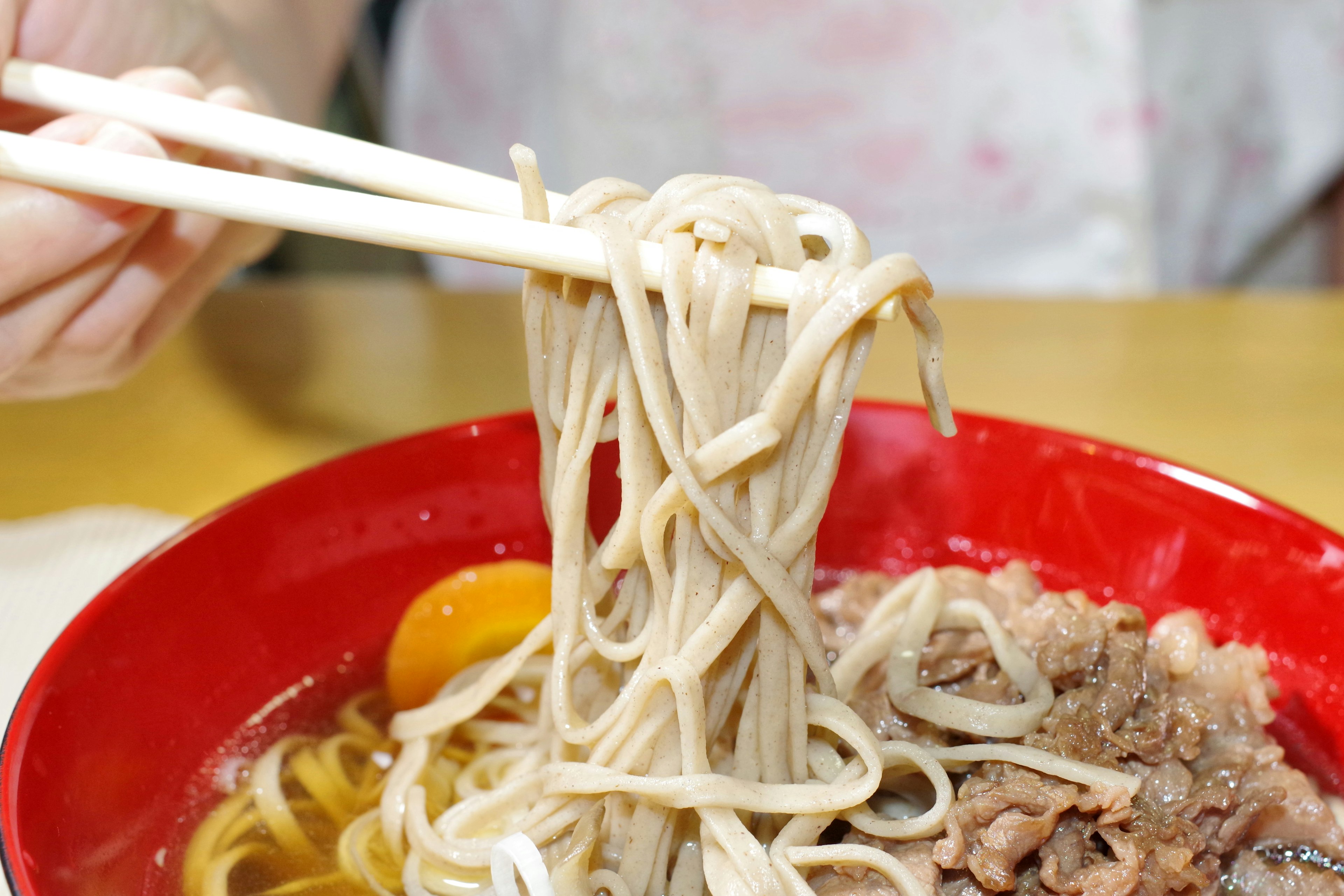 A hand lifting soba noodles from a red bowl