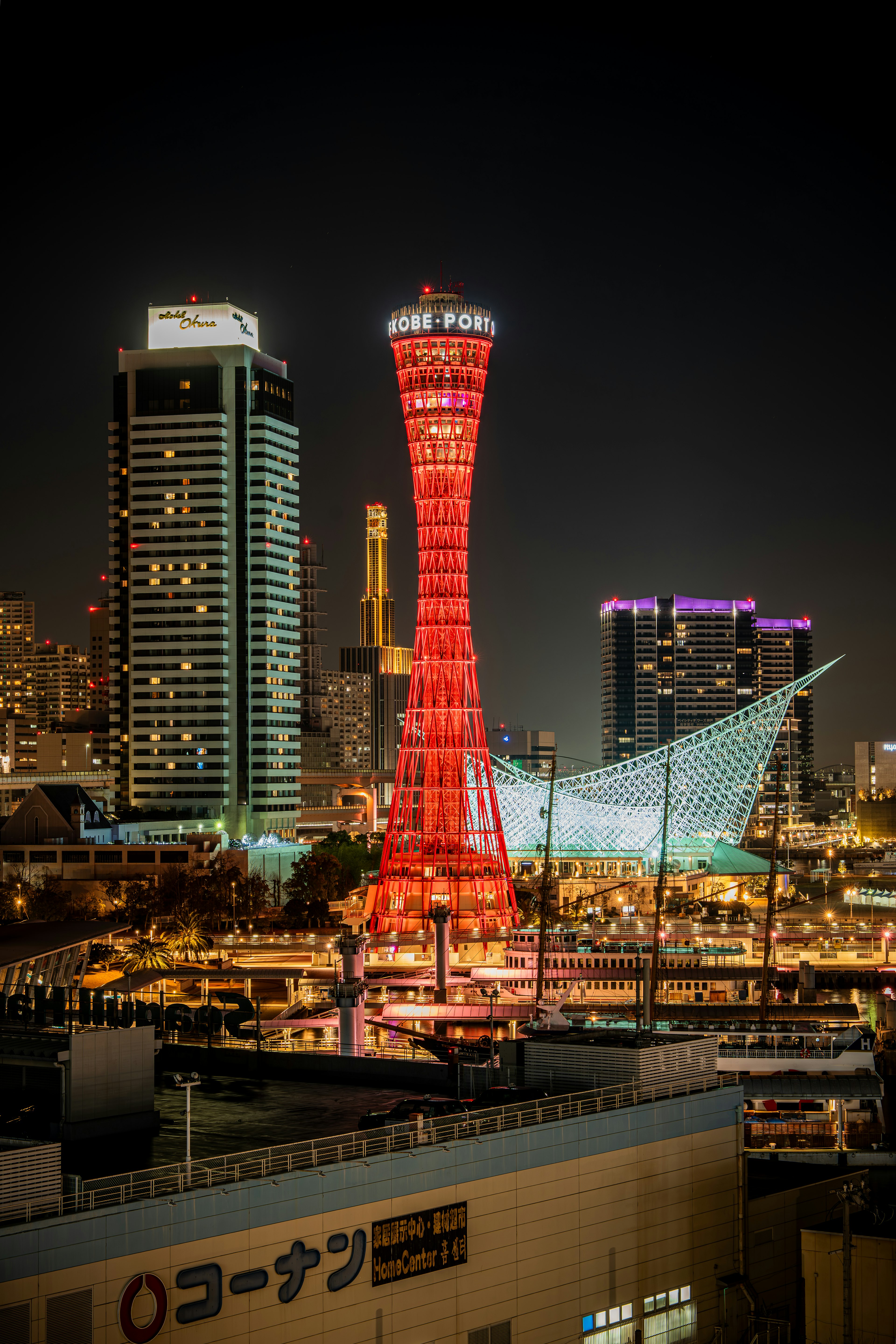 หอคอยท่าเรือโกเบสีน้ำเงินที่สว่างไสวในเวลากลางคืนพร้อมอาคารสมัยใหม่ในพื้นหลัง