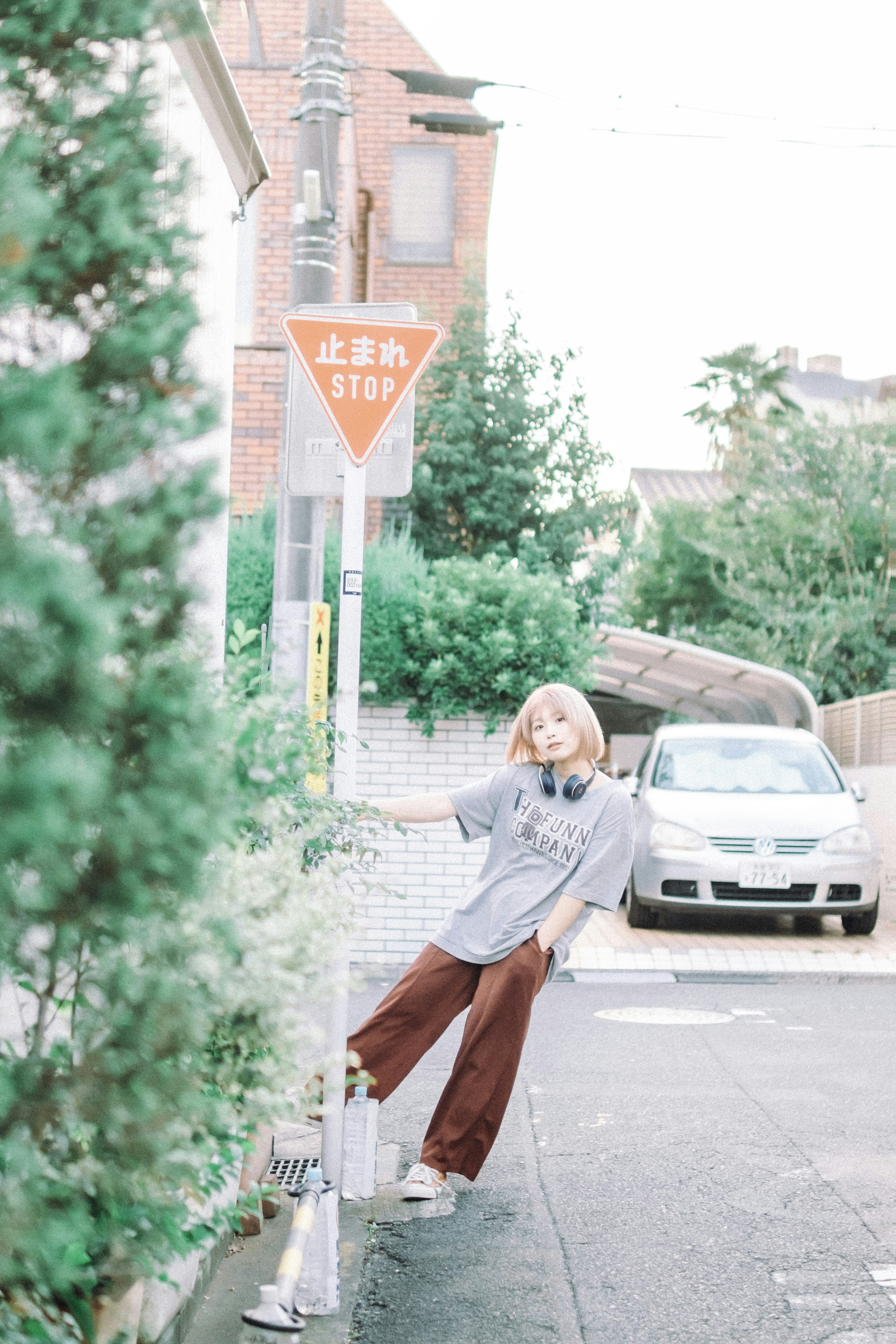 街角でポーズをとる女性　グレーのトップスと茶色のパンツ　交通標識と車が背景