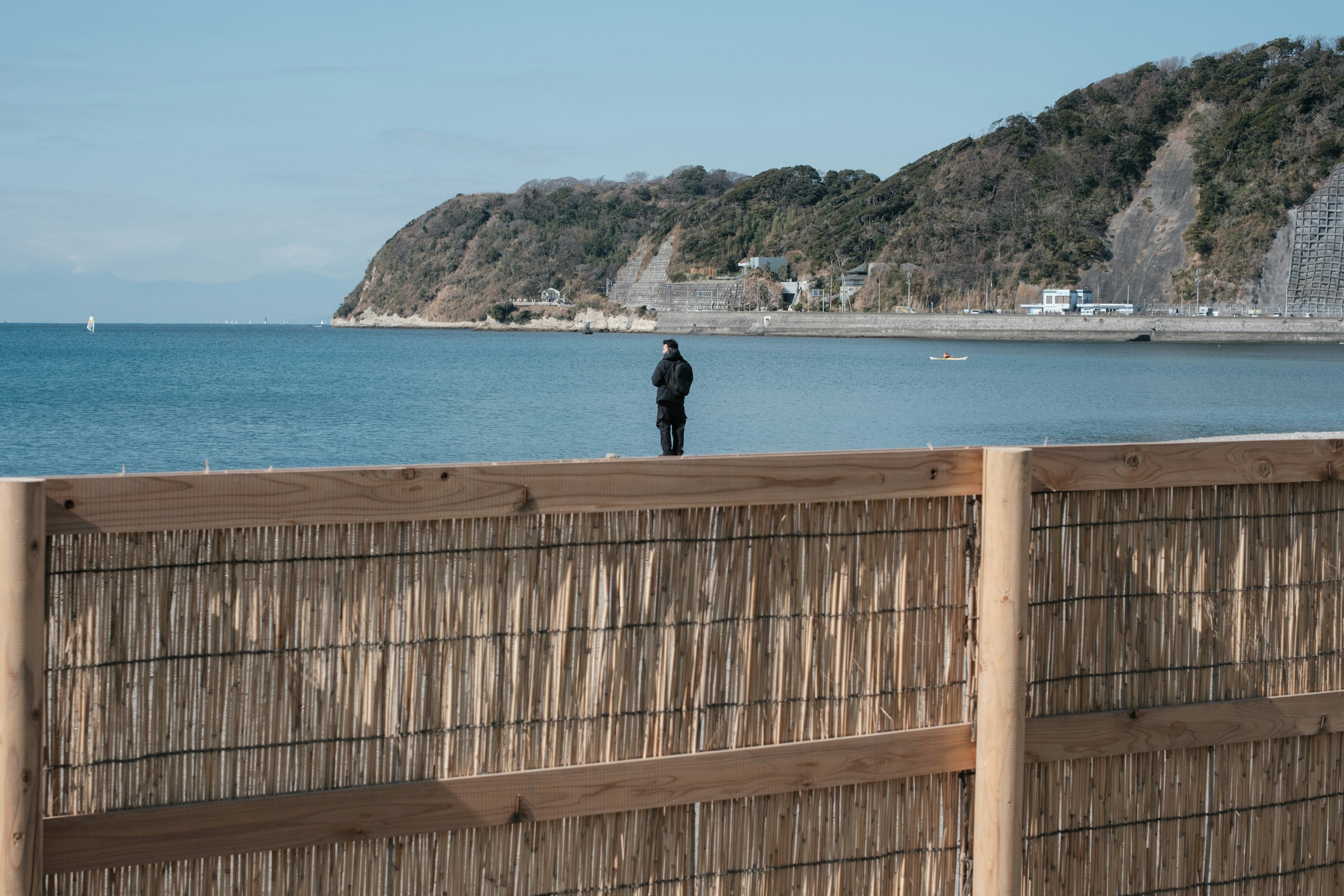 Person standing by the sea with a mountain backdrop and bamboo fence