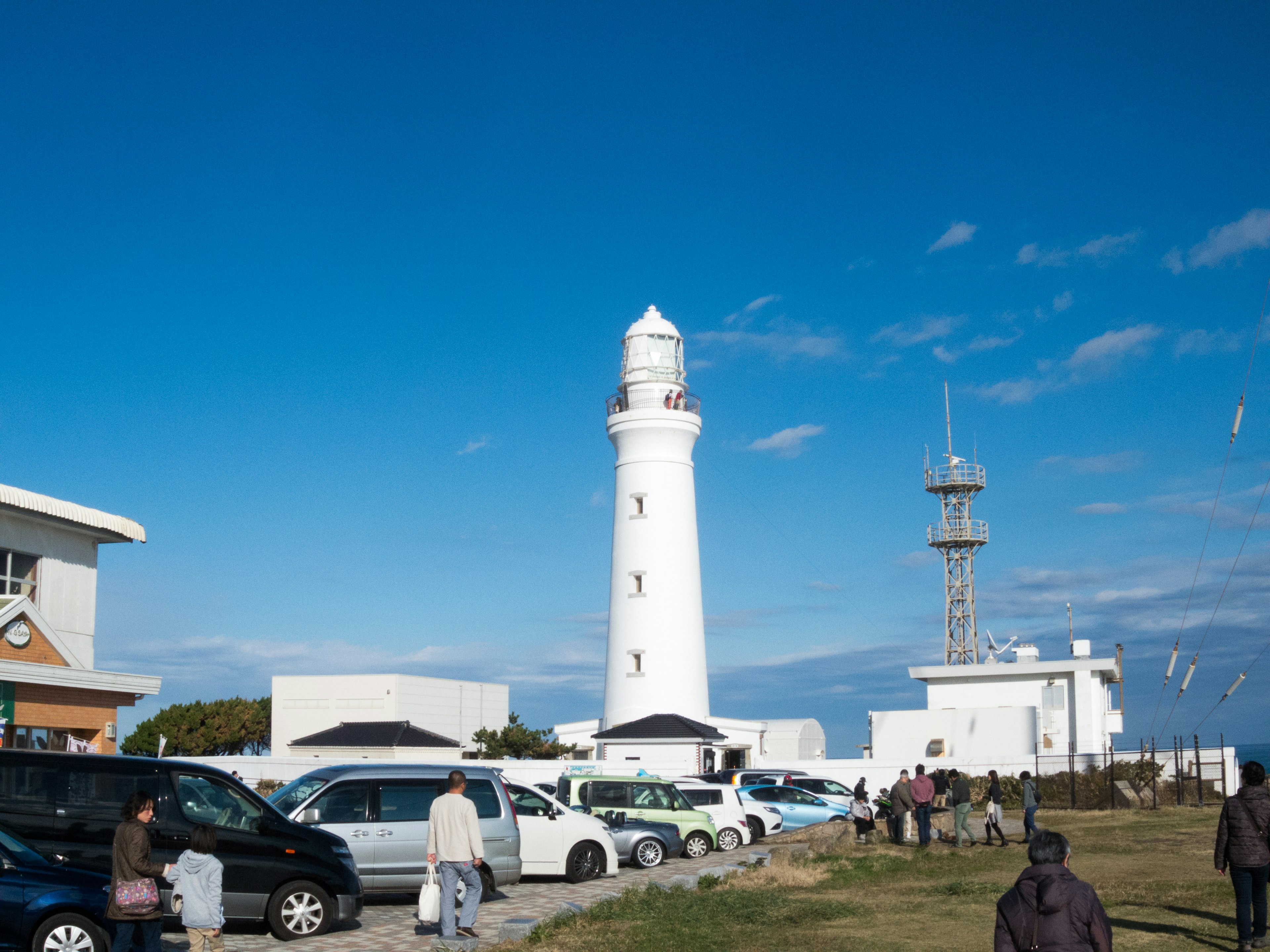 Un faro bianco contro un cielo blu con persone e auto in primo piano