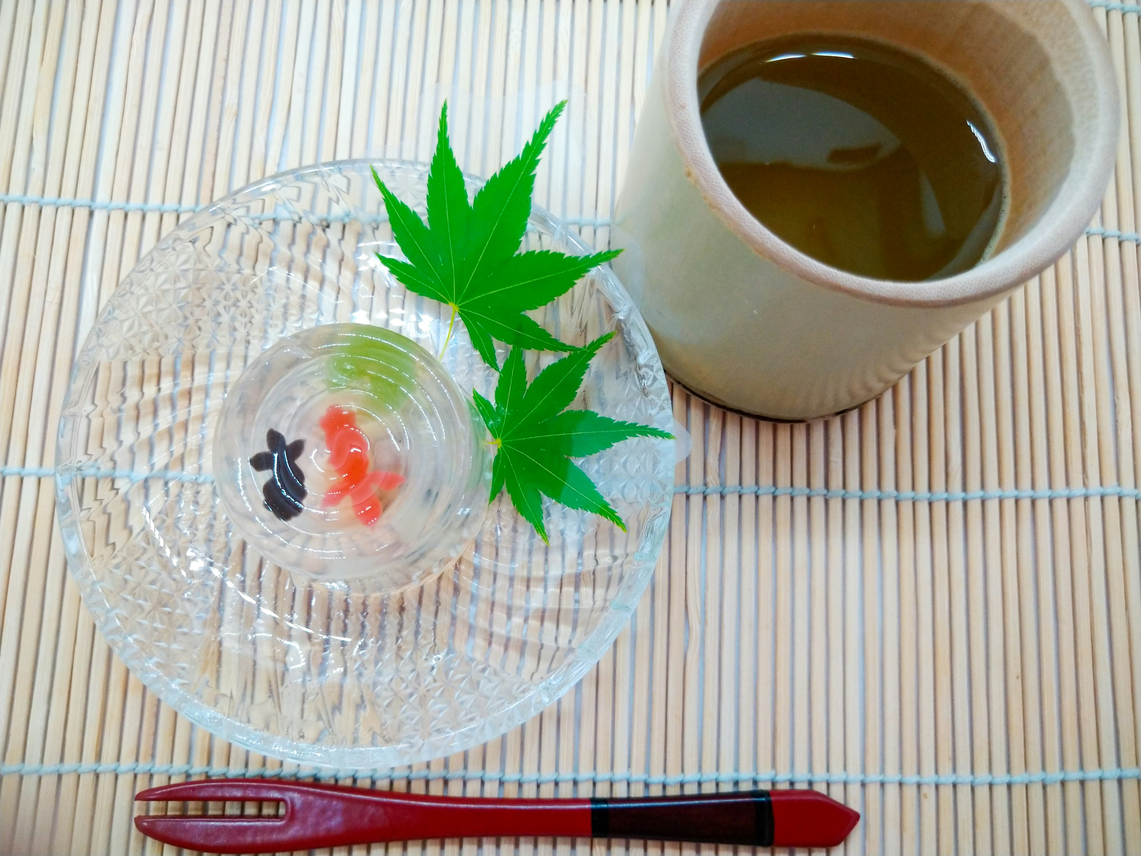 Ein klarer Teller mit traditionellen japanischen Süßigkeiten garniert mit grünen Ahornblättern und einer Tasse grünem Tee