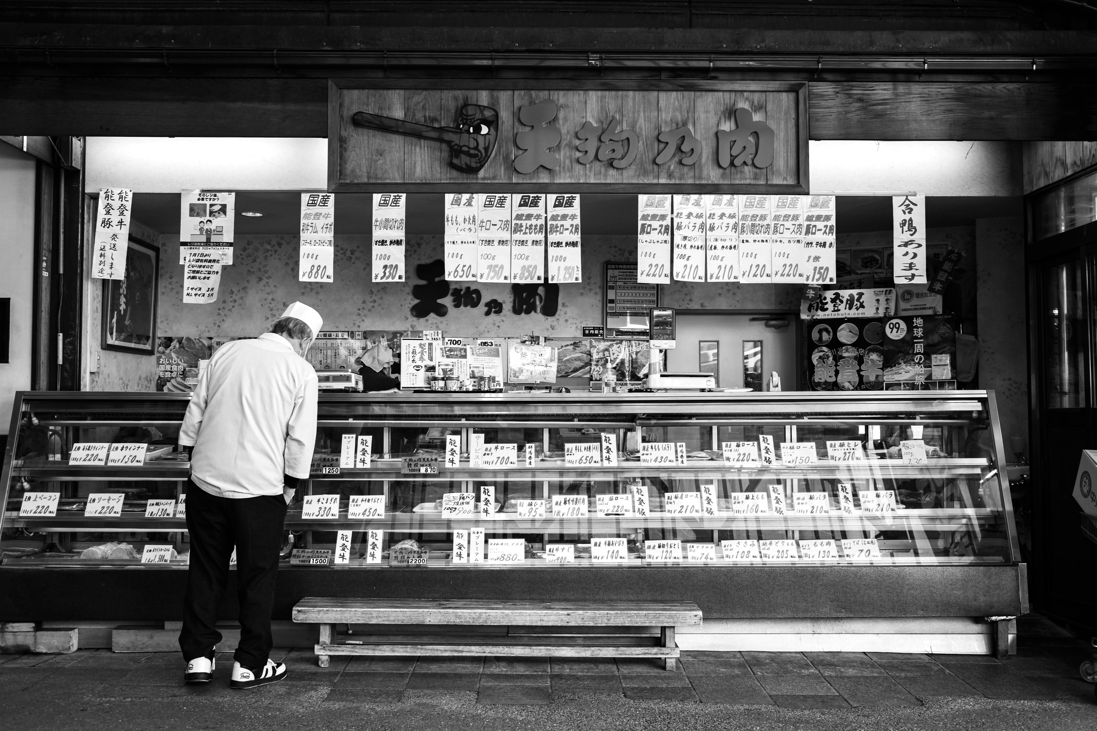 A man in white clothing standing in front of a traditional shop displaying various products