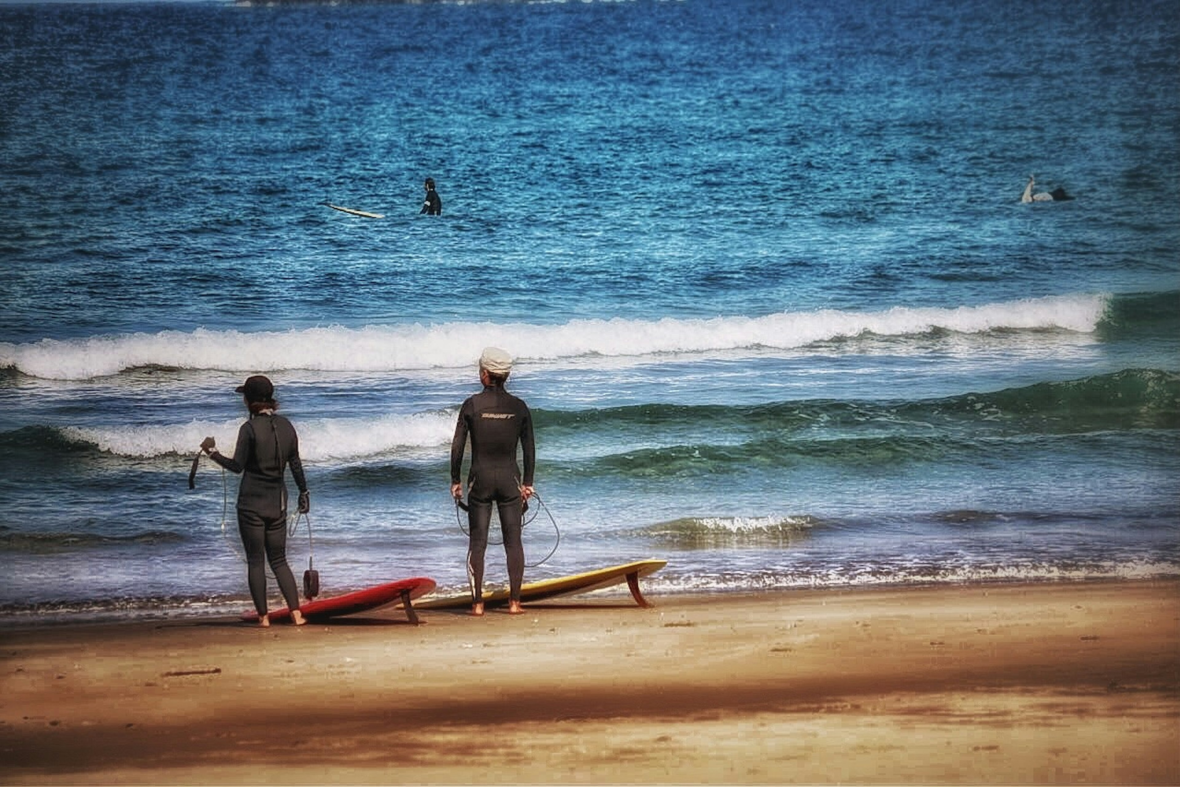 兩名衝浪者站在沙灘上，手持衝浪板，凝視著海浪