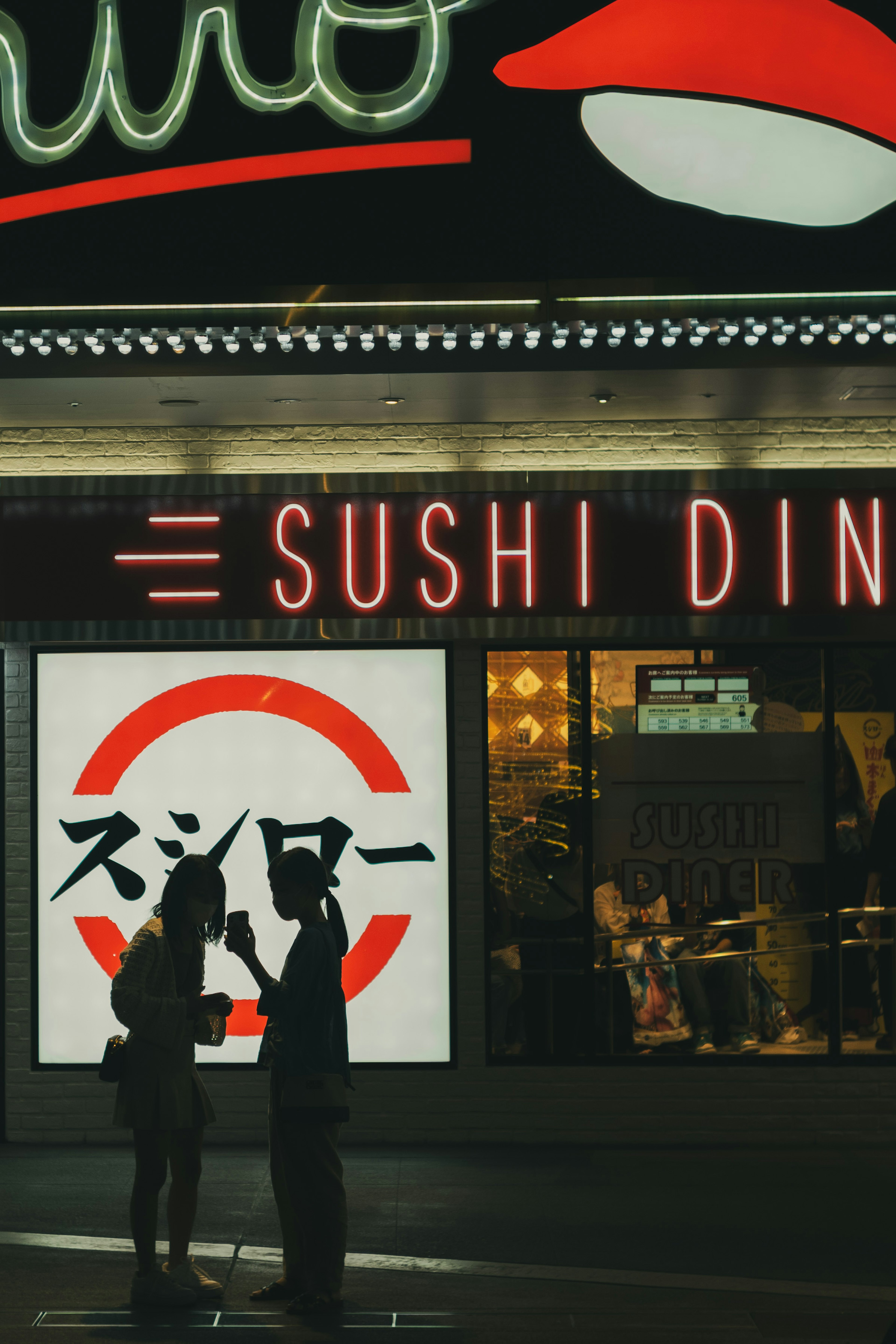 Silhouettes of two people conversing in front of a sushi restaurant with neon sign