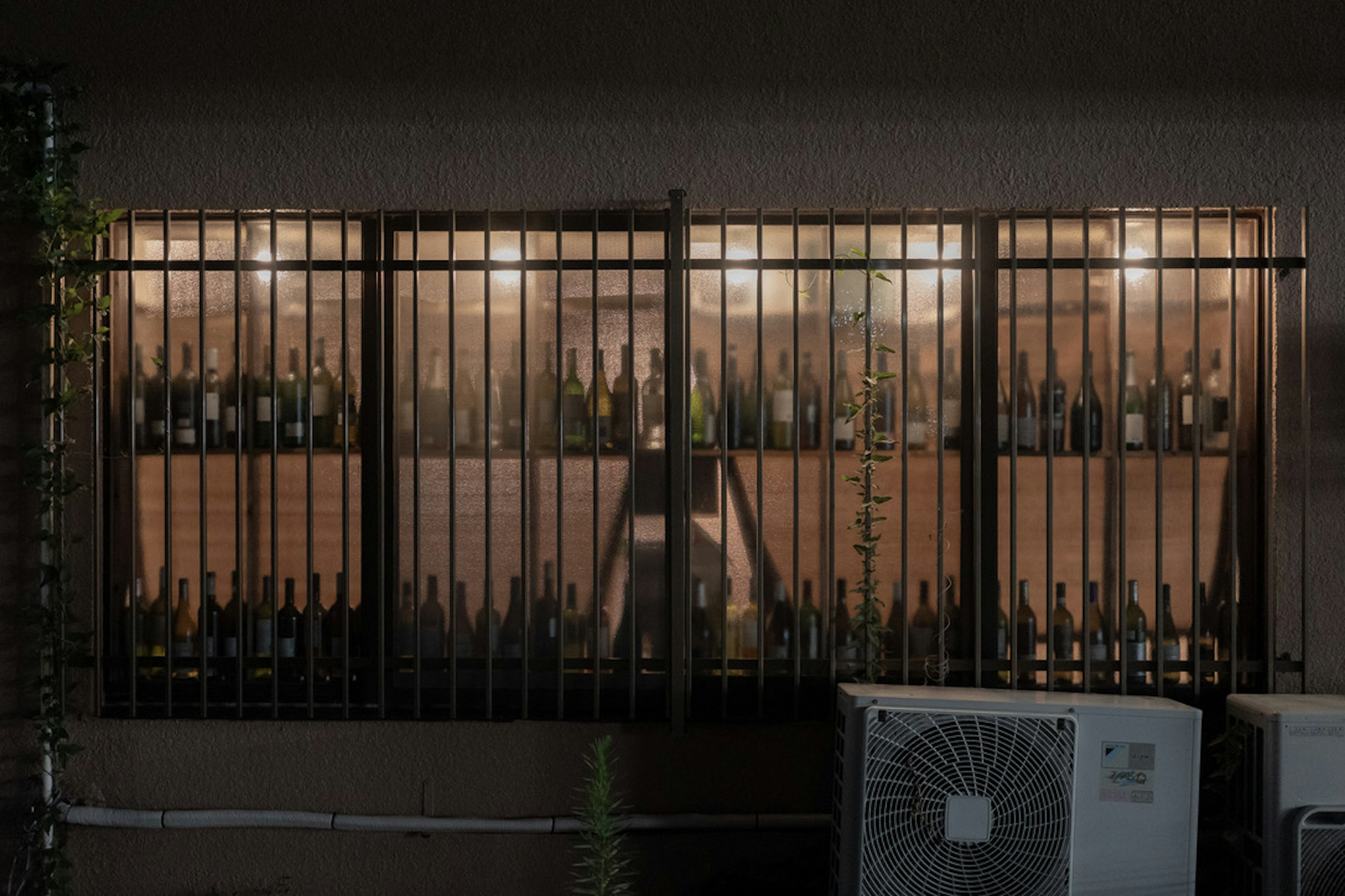 Night scene featuring a barred window displaying various bottles and warm lighting