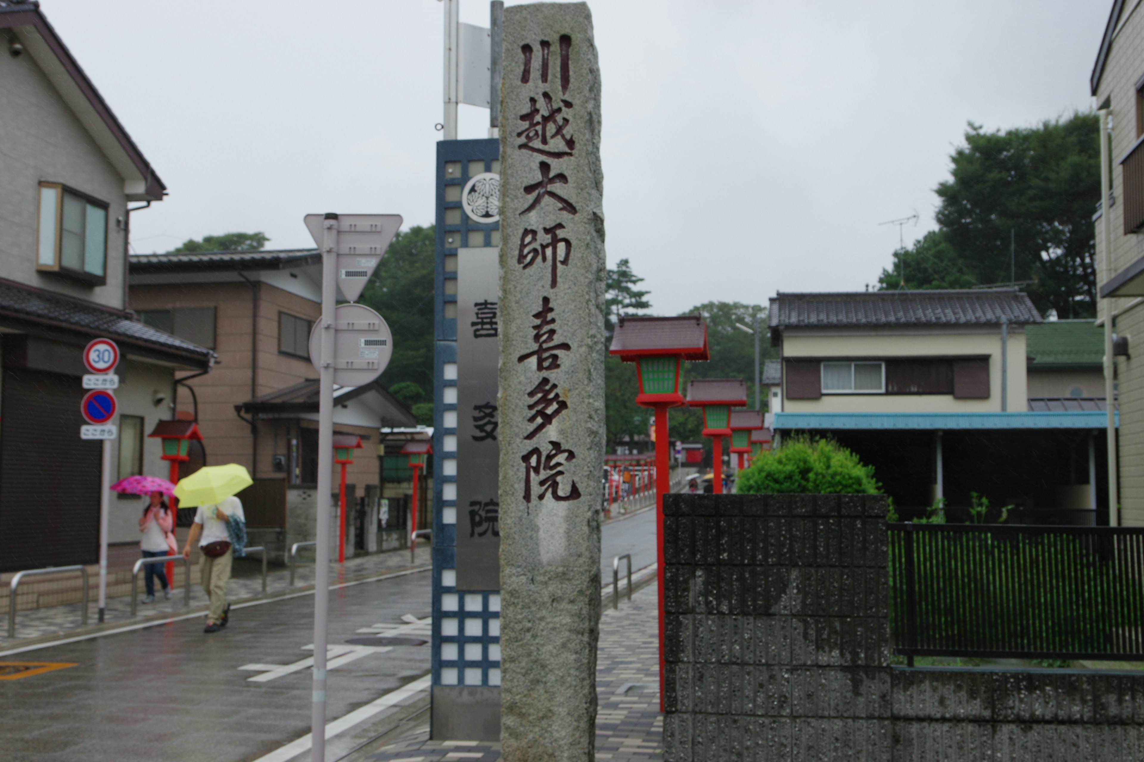 Steinmonument von Kawasuyo Daishi mit umliegender Straßenszene an einem regnerischen Tag