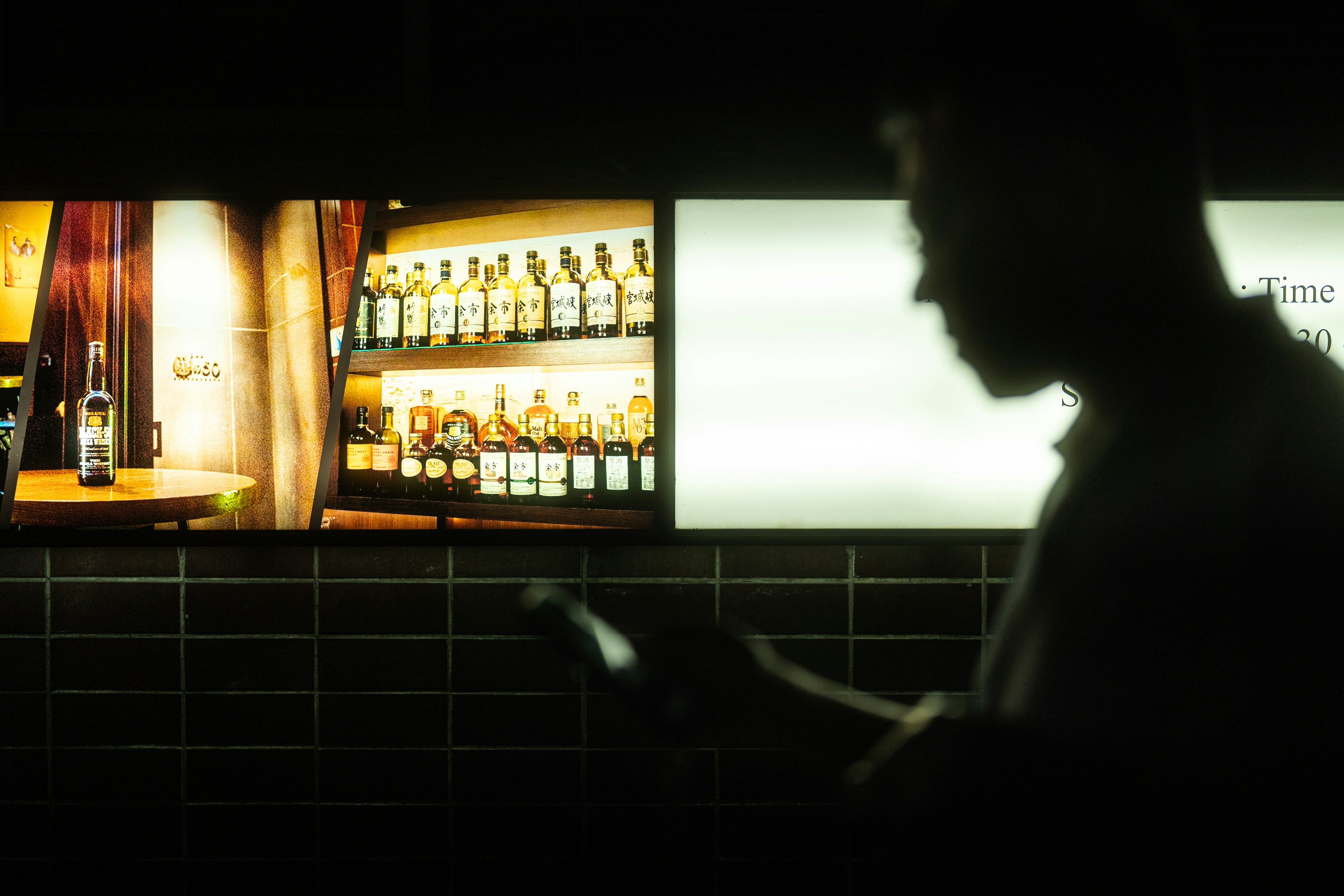 Silhouette di un uomo che tiene uno smartphone in un bar poco illuminato con scaffali di bottiglie sullo sfondo