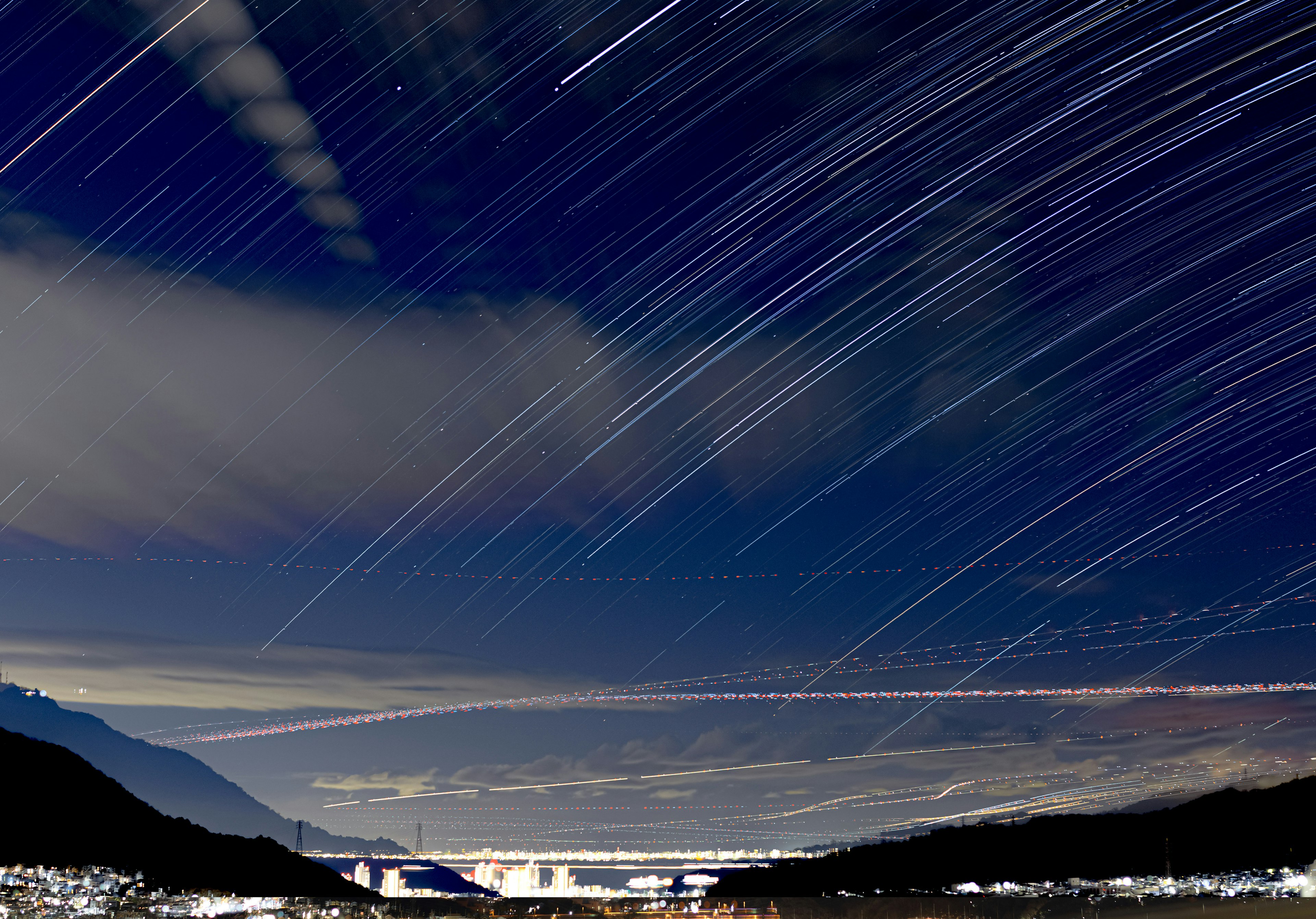 Night sky with star trails and clouds mountains in the background visible lights below