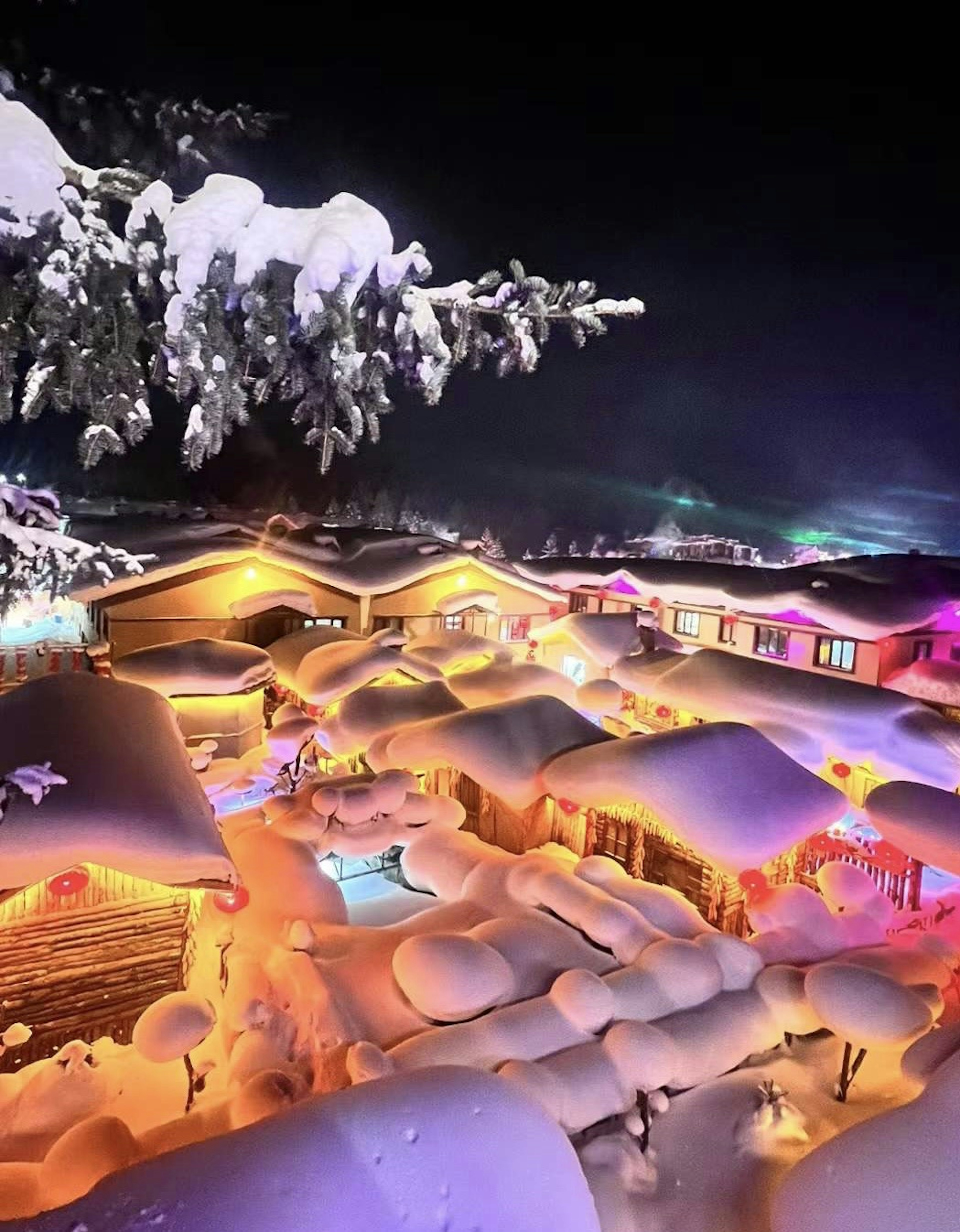 Snow-covered night scene with buildings illuminated by warm lights and a pool