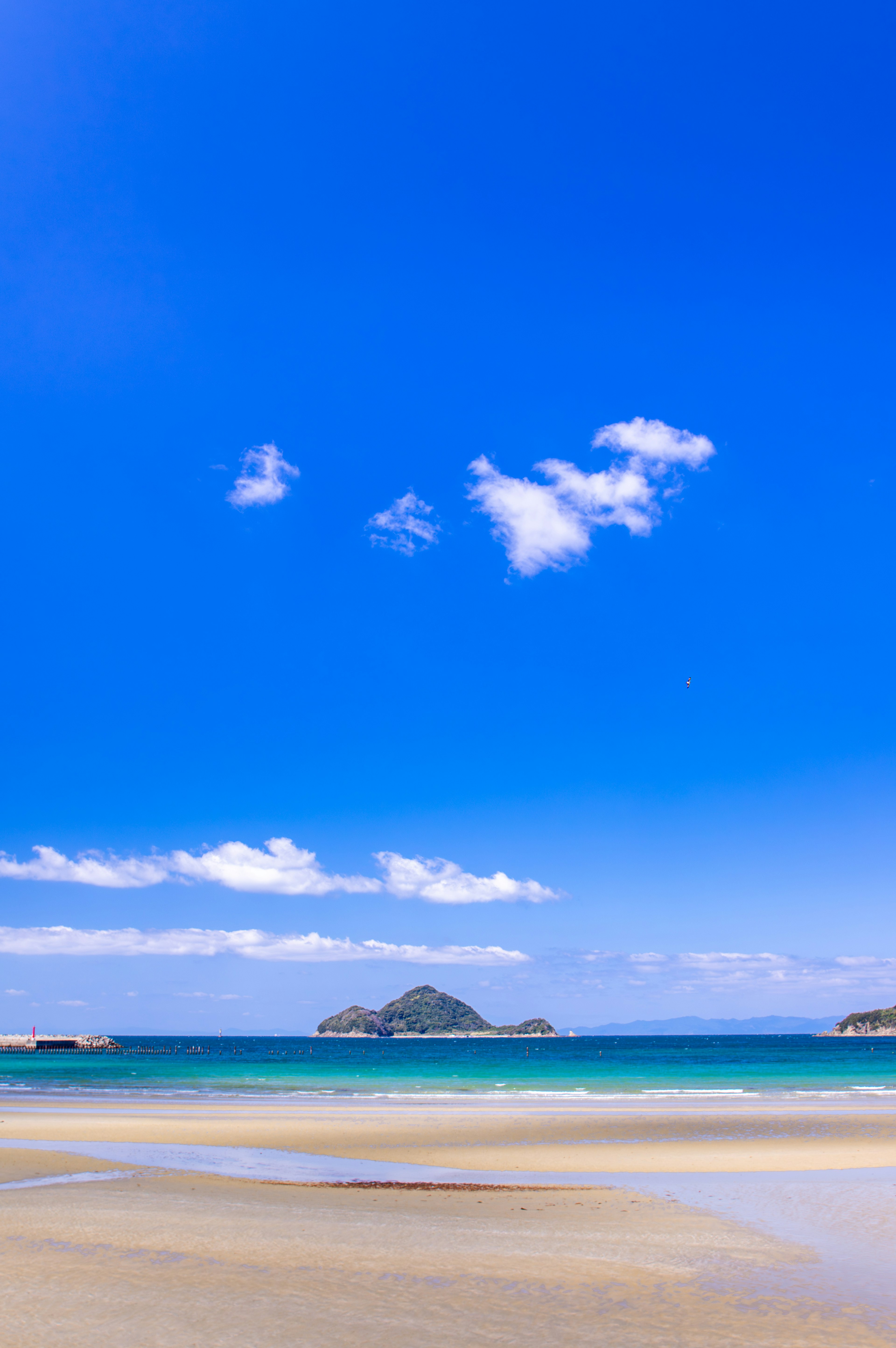 Eine malerische Strandansicht mit einem hellblauen Himmel und weißen Wolken über einem ruhigen Meer und einem sandigen Ufer