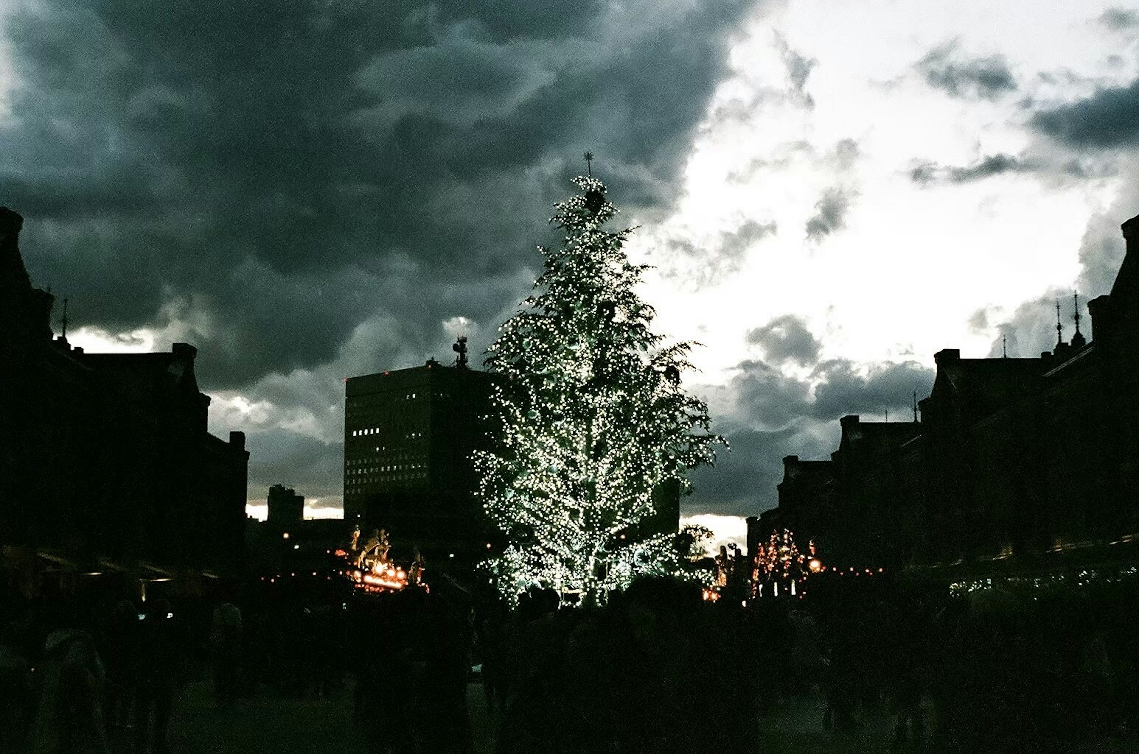 暗い雲に覆われた空の下に立つ輝くクリスマスツリーと周囲の人々